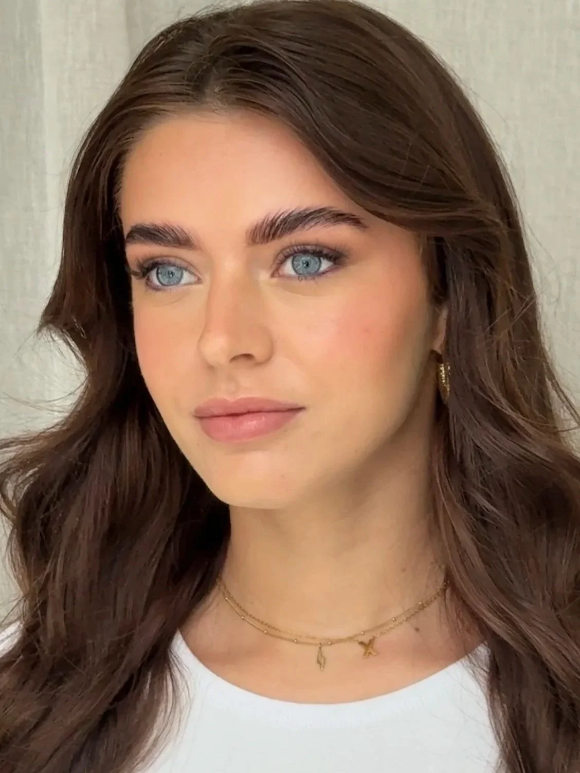 Close-up of a young woman with long, wavy brown hair and blue eyes, wearing subtle makeup, gold hoop earrings, a delicate gold necklace with small charms, and a white top.