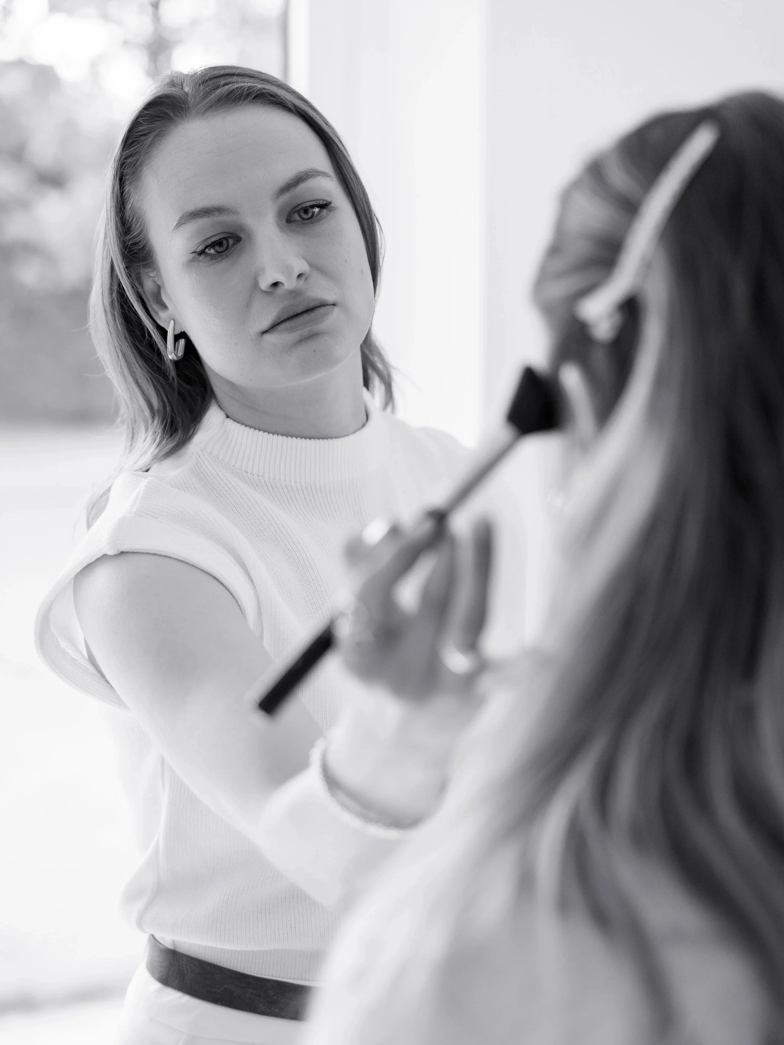 A woman with short hair and earrings applies makeup or cosmetics to another woman holding a makeup brush, in a well-lit room.