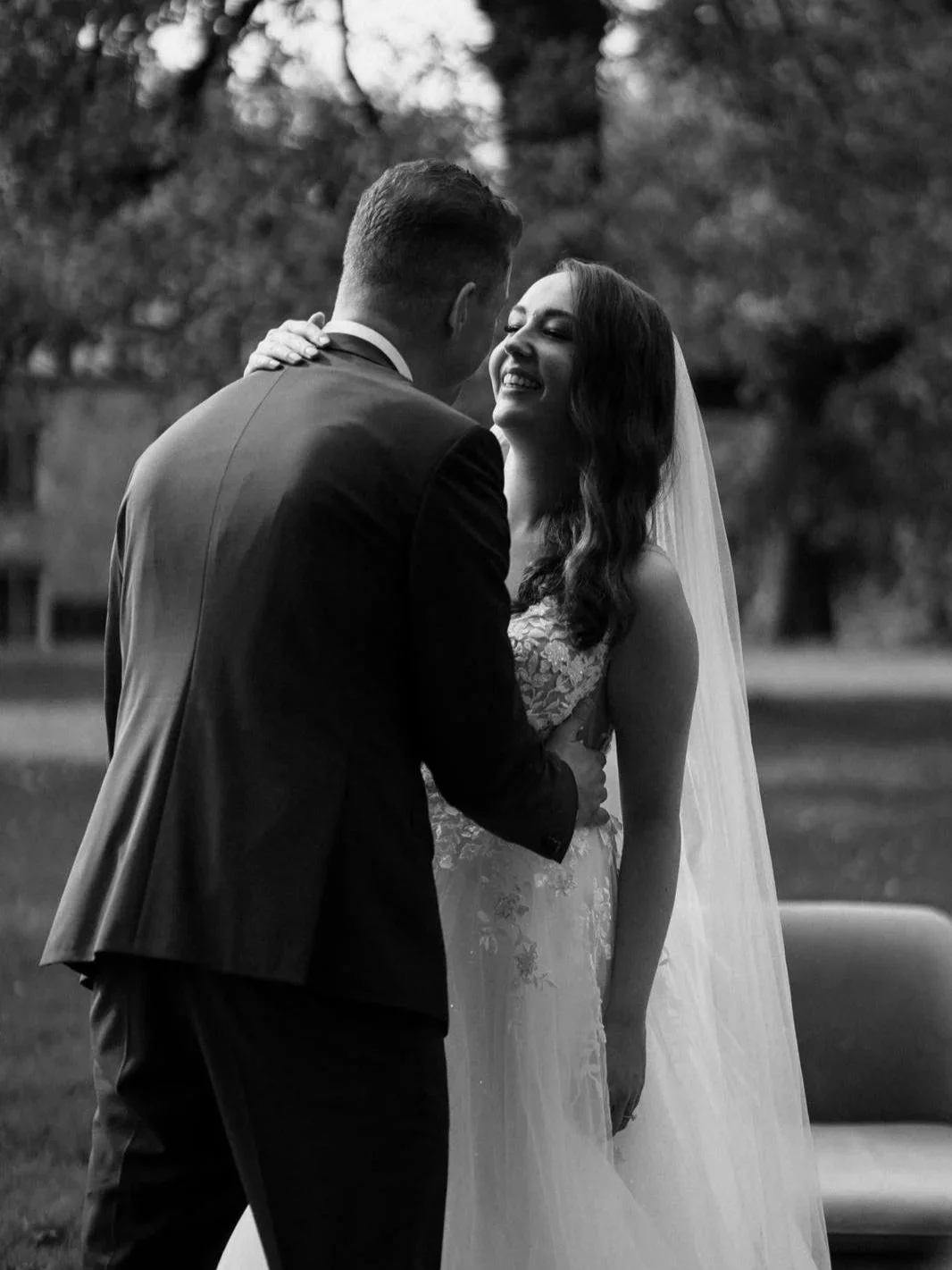 A black and white photo of a couple on their wedding day, smiling and embracing outdoors with trees in the background.