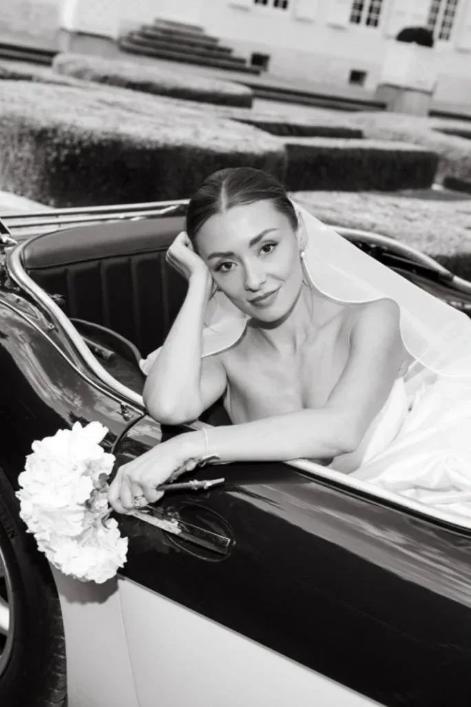 A woman in a wedding dress sitting in a convertible car holding a bouquet of flowers.