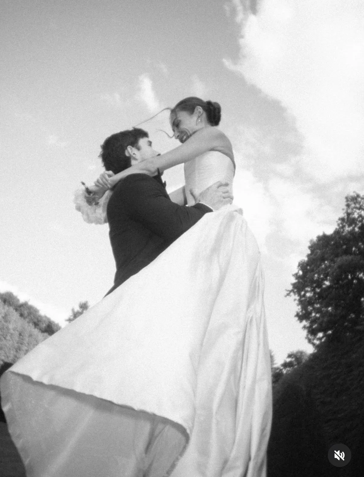 A couple dancing outdoors in wedding attire, with the woman lifted in the air by the man, against a background of trees and a cloudy sky.