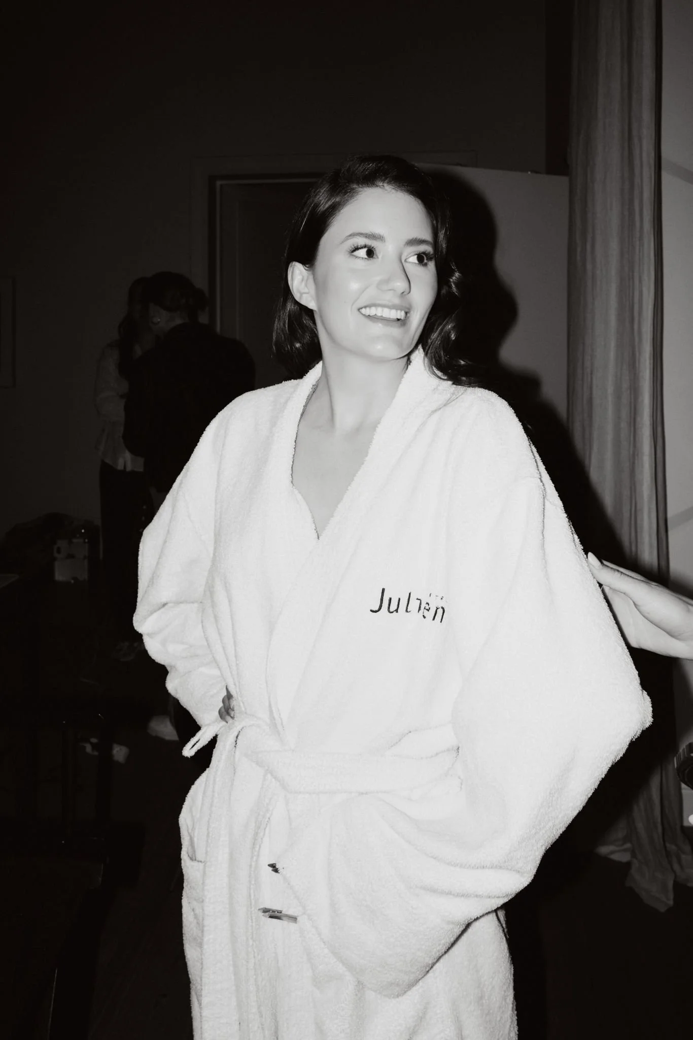 A woman with dark shoulder-length hair in a white bathrobe with the name 'Julien' embroidered on it, standing indoors. She is smiling and looking to the side.