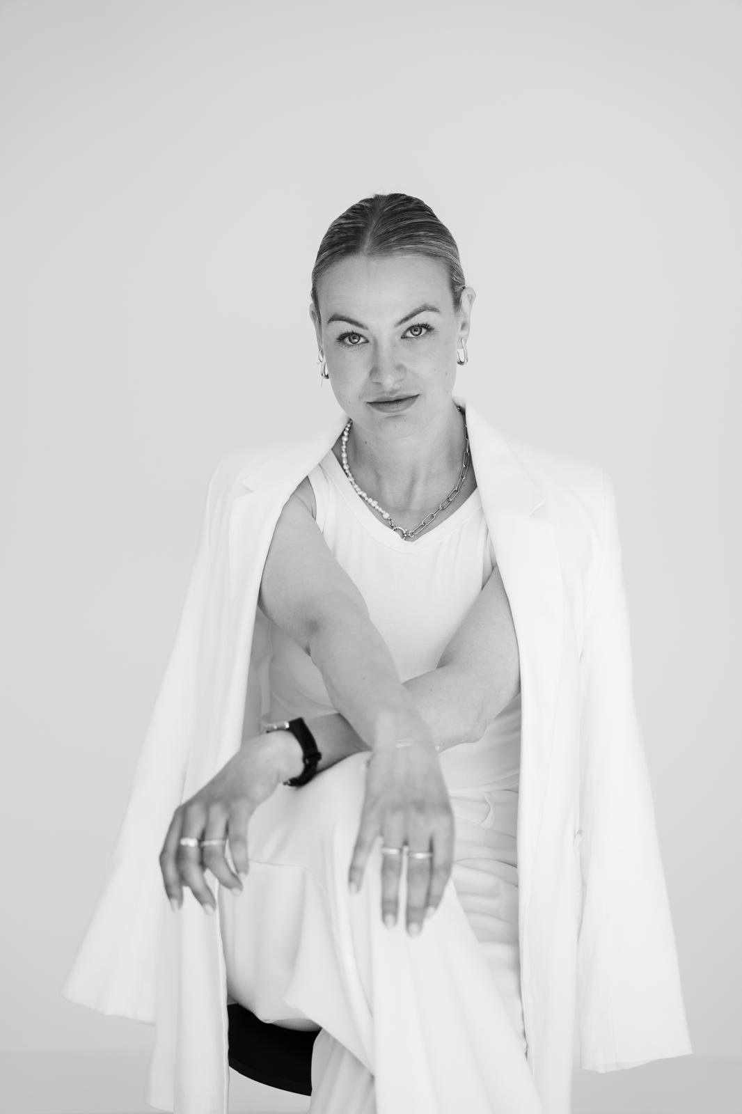 Black and white portrait of a woman sitting on a chair with her arm extended forward, looking directly at the camera with a confident expression, wearing a white sleeveless top, a light-colored blazer draped over her shoulders, and jewelry including earrings, necklaces, rings, and a watch.