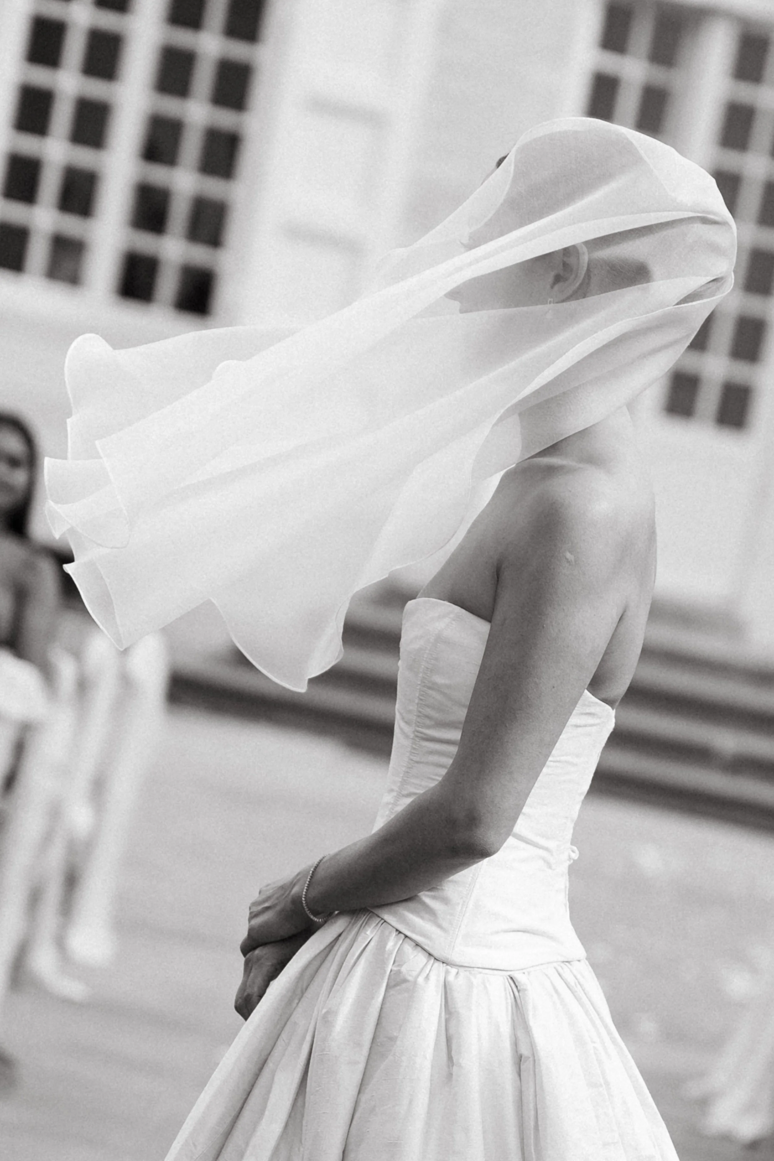 A woman in a strapless gown with a billowing veil on her head, standing outdoors with a blurred background.