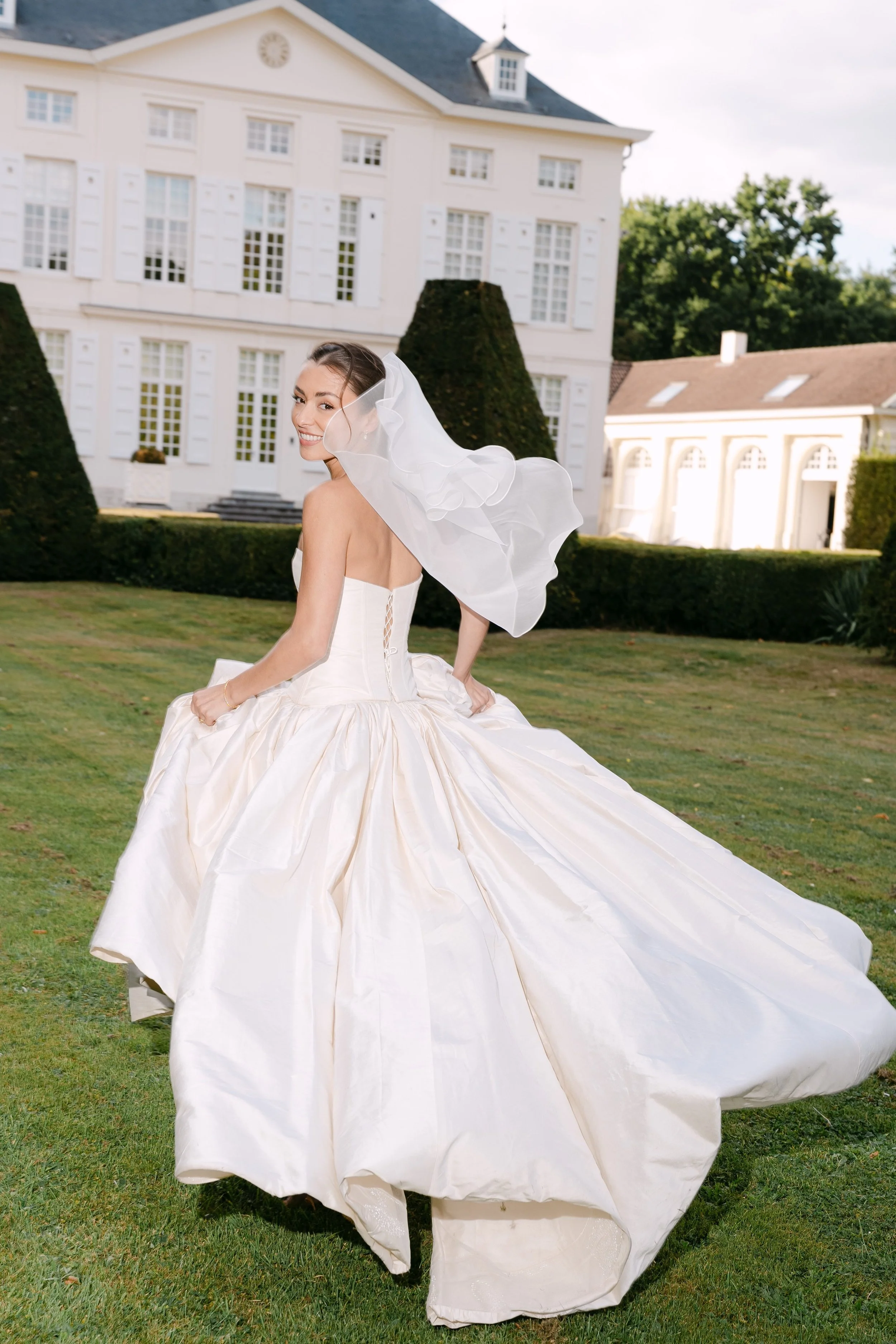 A smiling woman in a wedding dress with a veil, standing on a lawn in front of a large white house with shutters and a smaller building in the background.