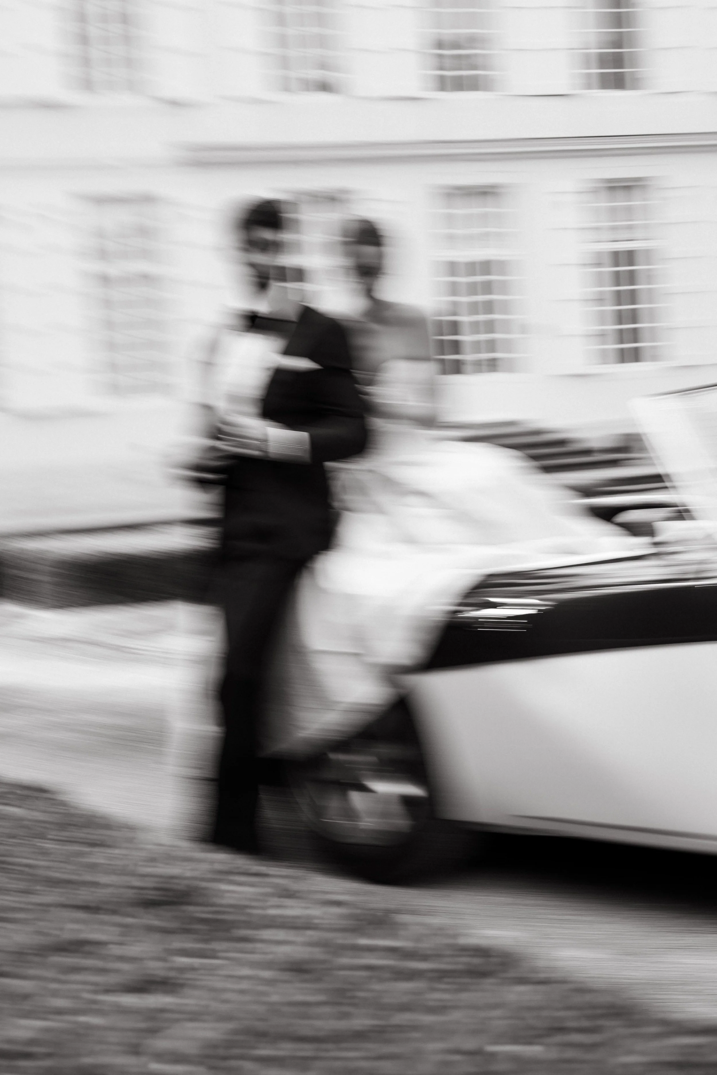 Blurry black-and-white photo of a man standing next to a vintage car outside a building with windows.