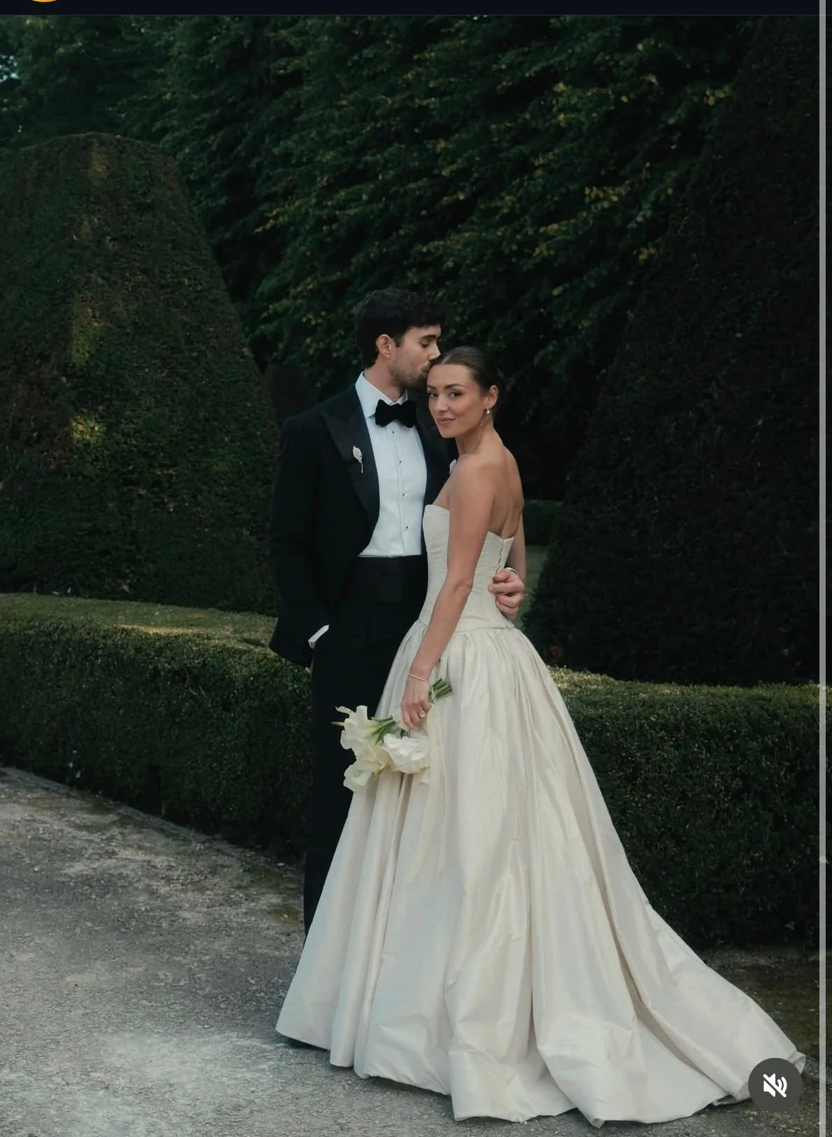 A bride and groom posing outdoors, the groom is wearing a black tuxedo and bowtie, and the bride is in a strapless white wedding gown holding a bouquet of white calla lilies.