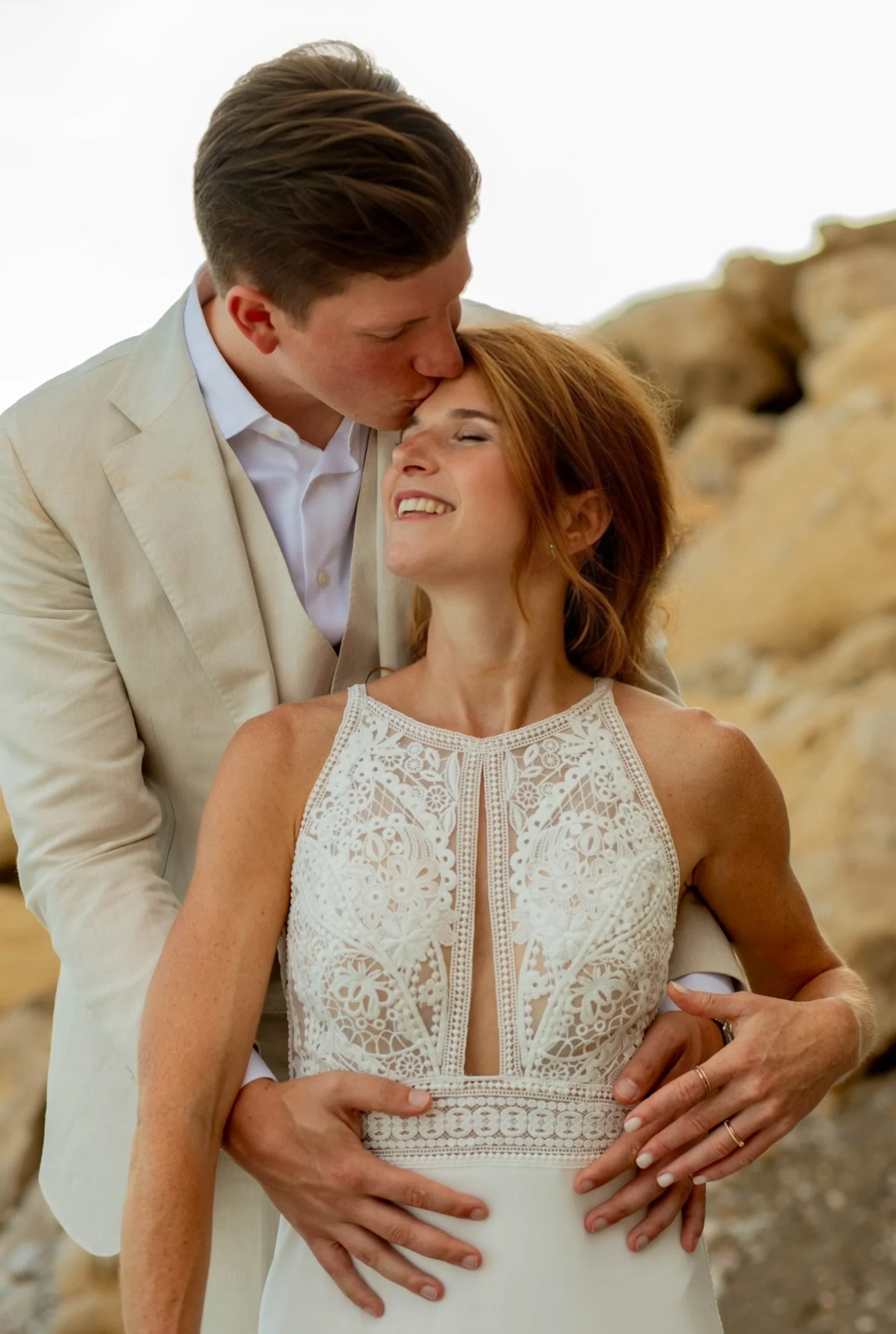 A man and woman at a beach, with the man kissing the woman's forehead. The woman is smiling with her eyes closed, wearing a white lace dress, and the man is dressed in a beige suit.