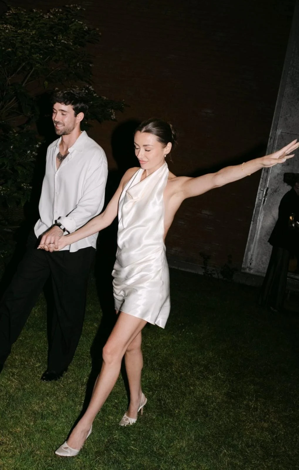 A woman in a silk dress and heels stretches her arms out as she walks with a man in a white shirt and black pants at night outdoors.