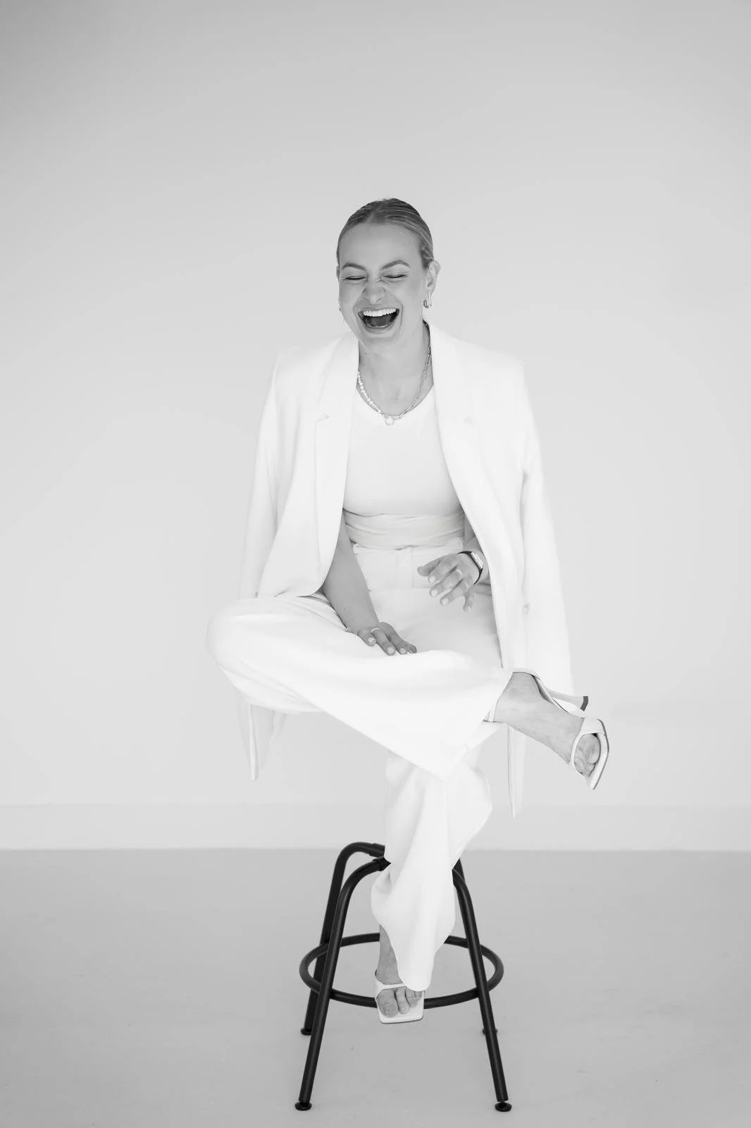 Black and white photo of a woman laughing, sitting on a stool with one leg crossed over the other, wearing a white suit and high heels.
