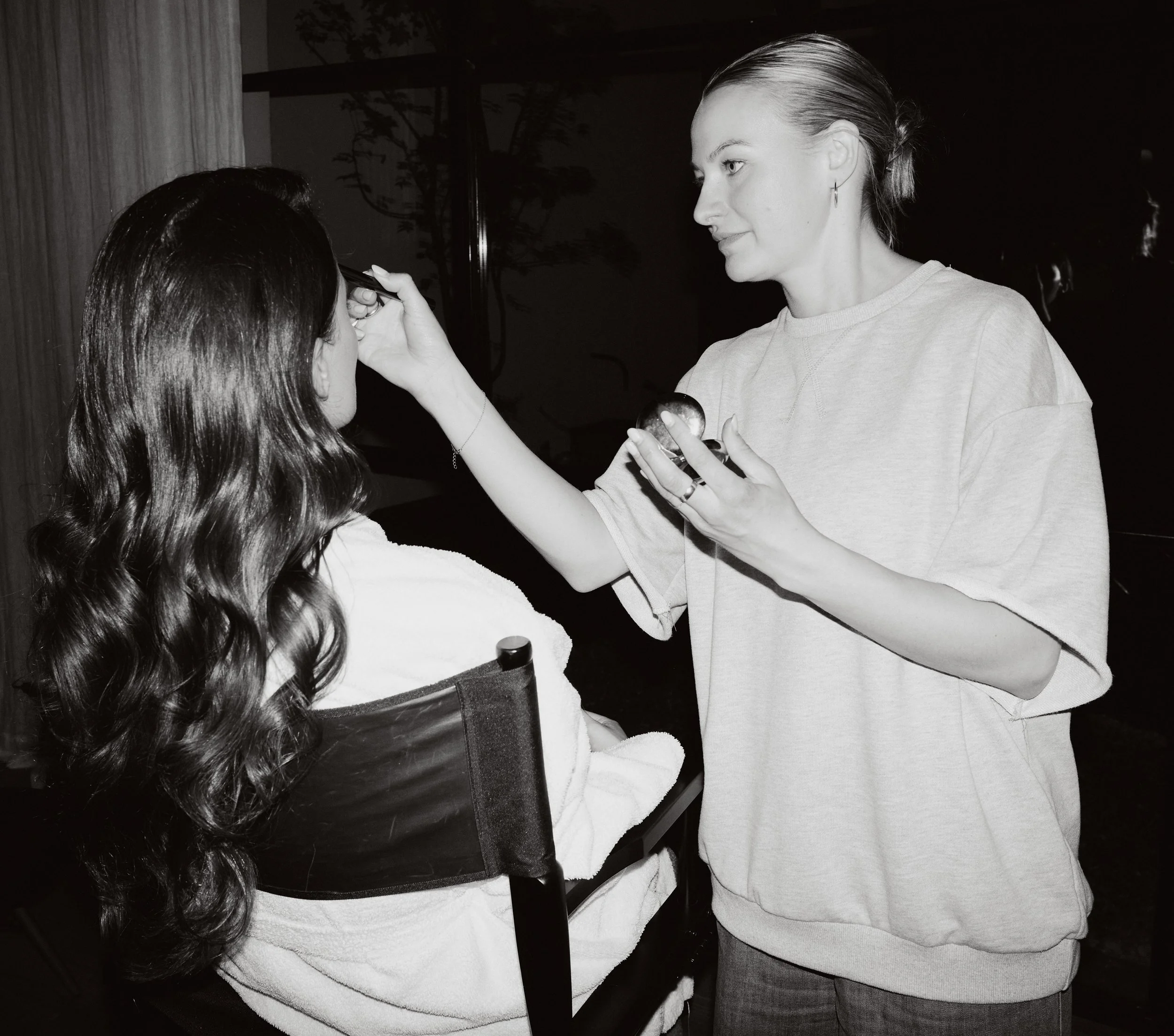 A woman with long wavy hair applying makeup to another woman sitting in a wheelchair at night.