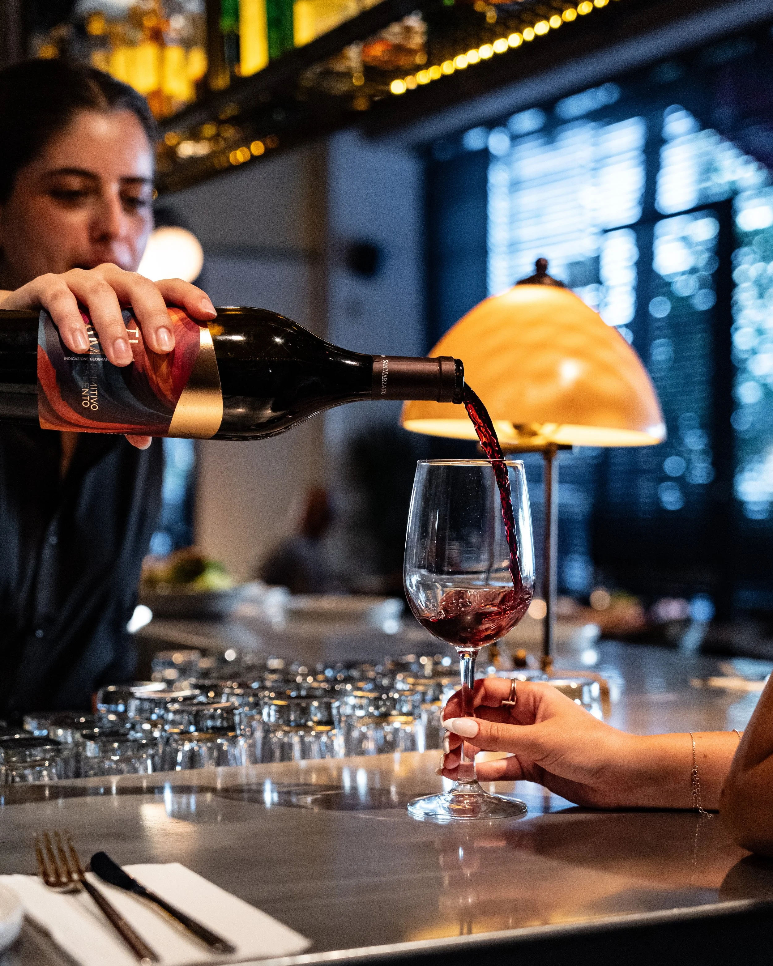 A person is pouring red wine into a wine glass at a restaurant or bar, with another person's hand holding the glass. There are glasses, a lamp, and other objects in the background.