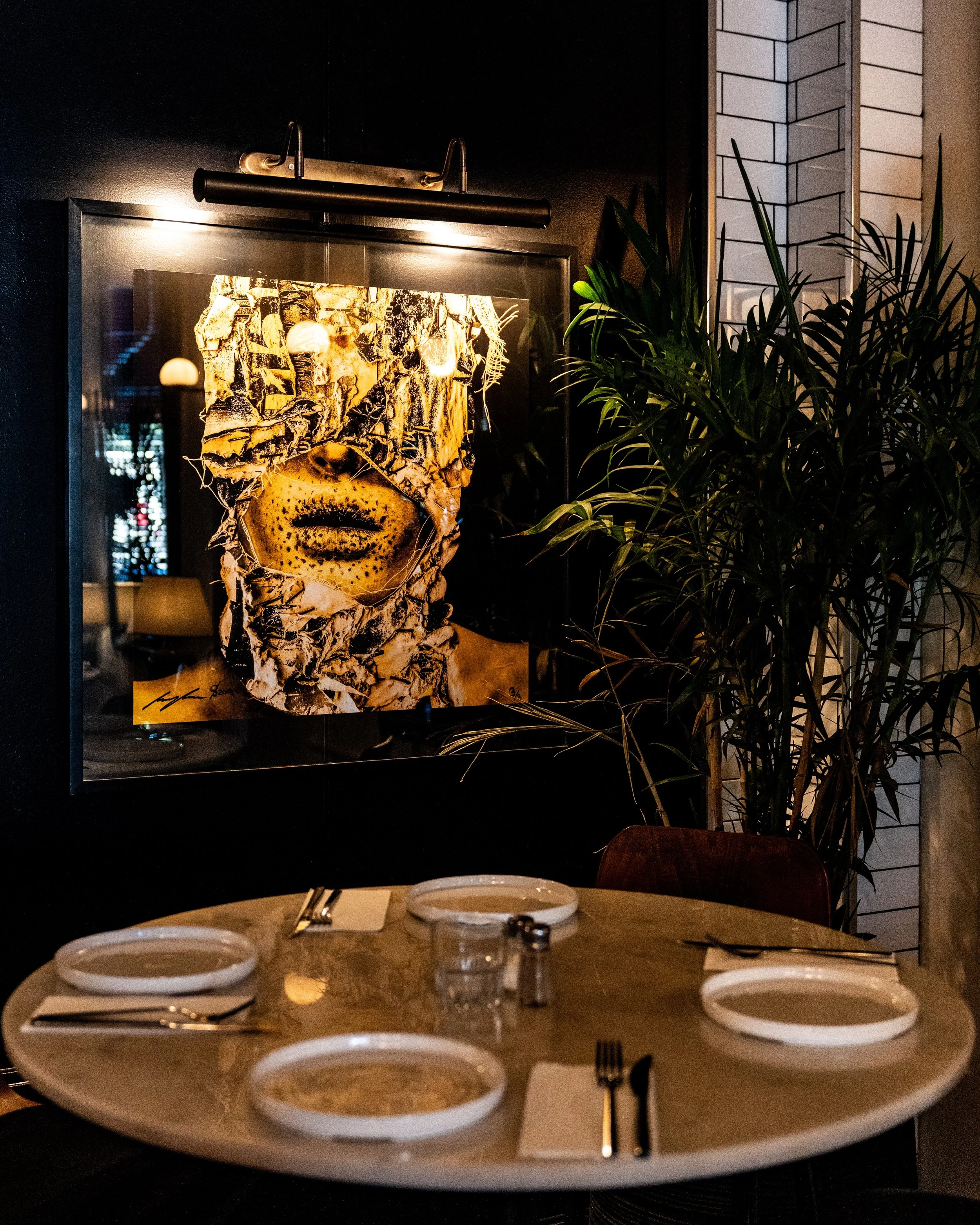 A round dining table set with four white plates, forks, and napkins in a dimly lit restaurant. Behind the table, a framed portrait of a woman's face is displayed on a dark wall, partially obscured by a large green plant.