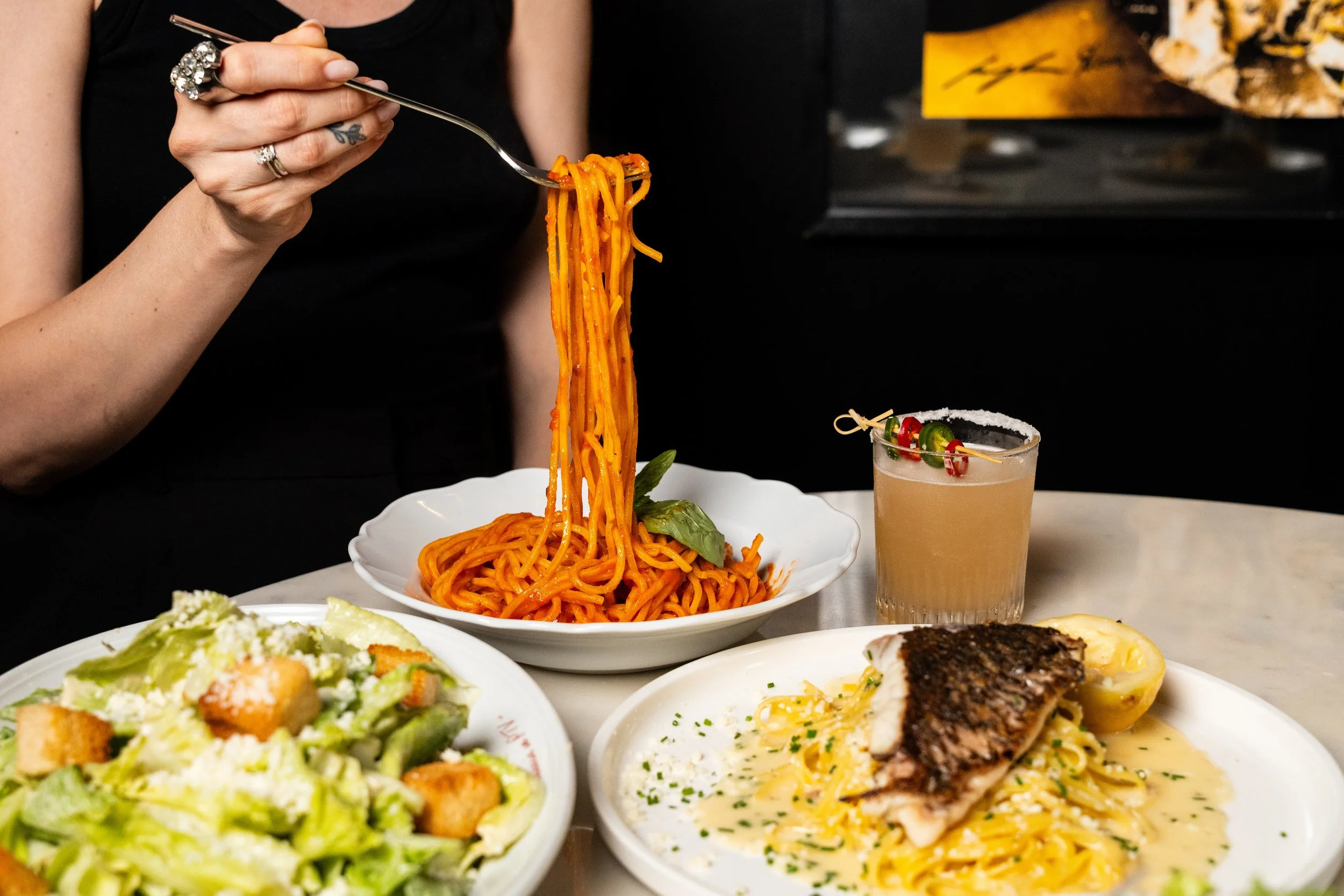 Person lifting a forkful of spaghetti from a plate of pasta with basil leaves, with a salad and a dish of fish with creamy sauce on the table, along with a cocktail garnished with cherries and jalapenos