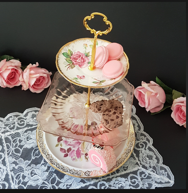 Three-tiered porcelain serving stand with pink macarons on top, chocolate chip cookies on middle tier, and a wrapped pink and white rolled sweet on the bottom. The stand is decorated with floral patterns and is placed on a lace tablecloth surrounded by pink roses.