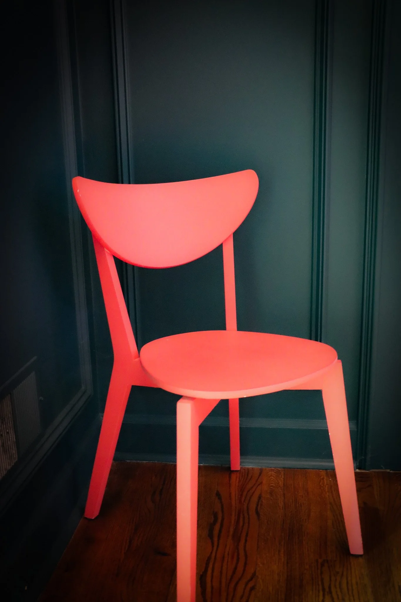A bright pink modern chair with a curved backrest and four legs, positioned in front of dark green paneled walls and wooden flooring.