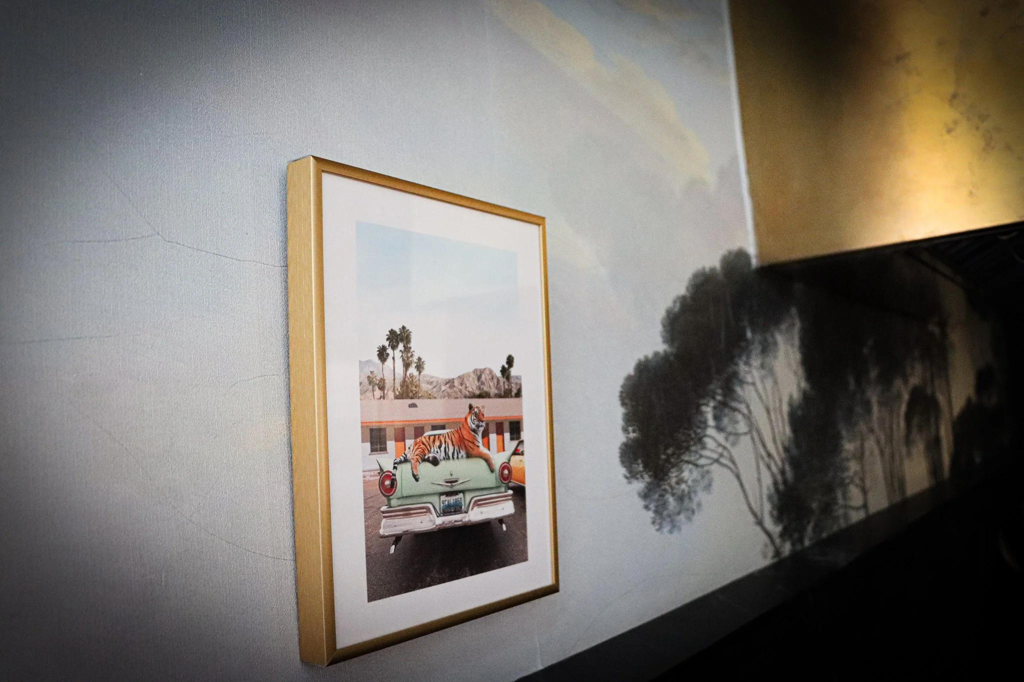 A framed photo on a wall showing a vintage car with a tiger lying on its trunk, with palm trees and mountains in the background.