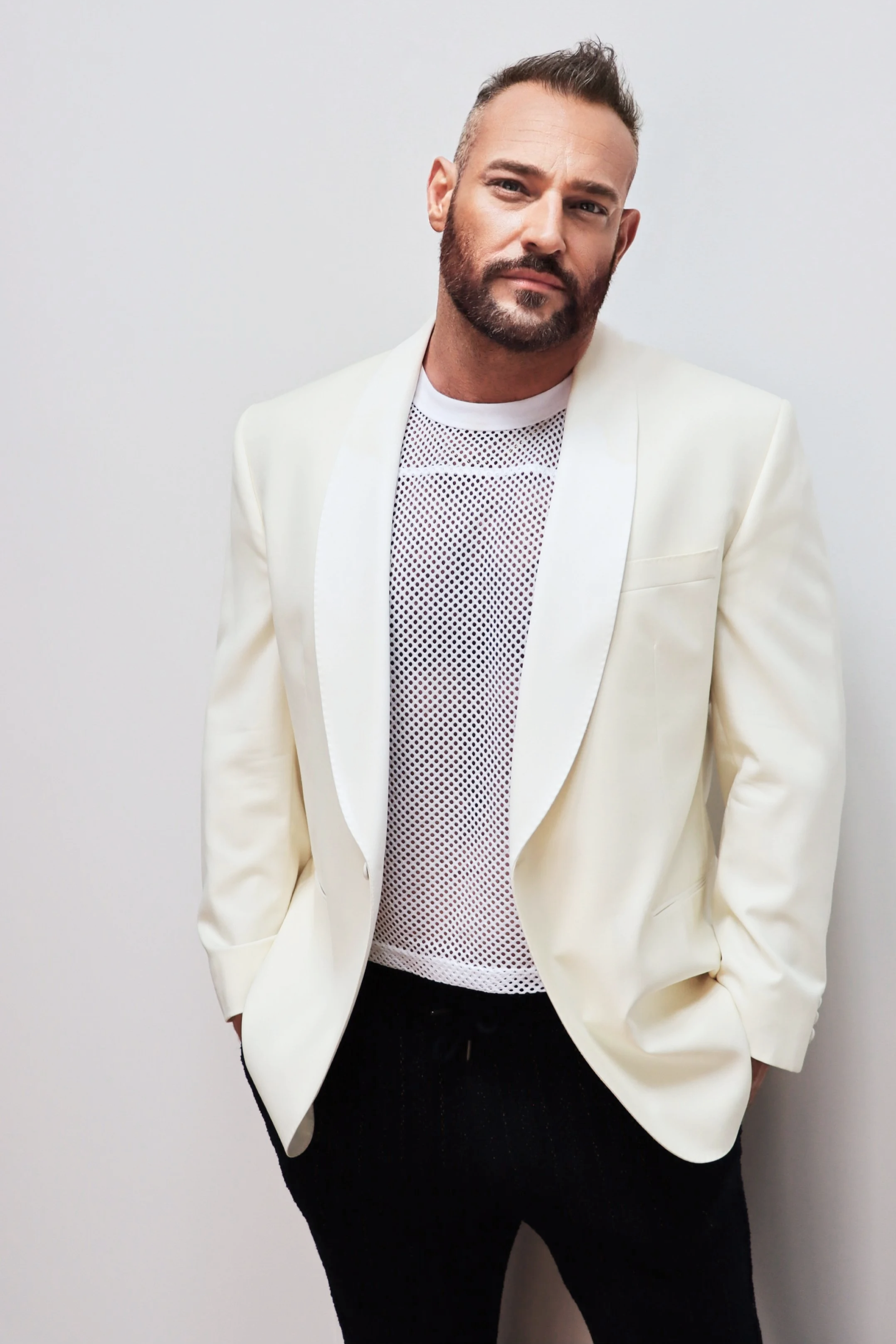 A man with a beard and styled hair wearing a white blazer, a white mesh shirt, and black pants, standing against a plain white wall.