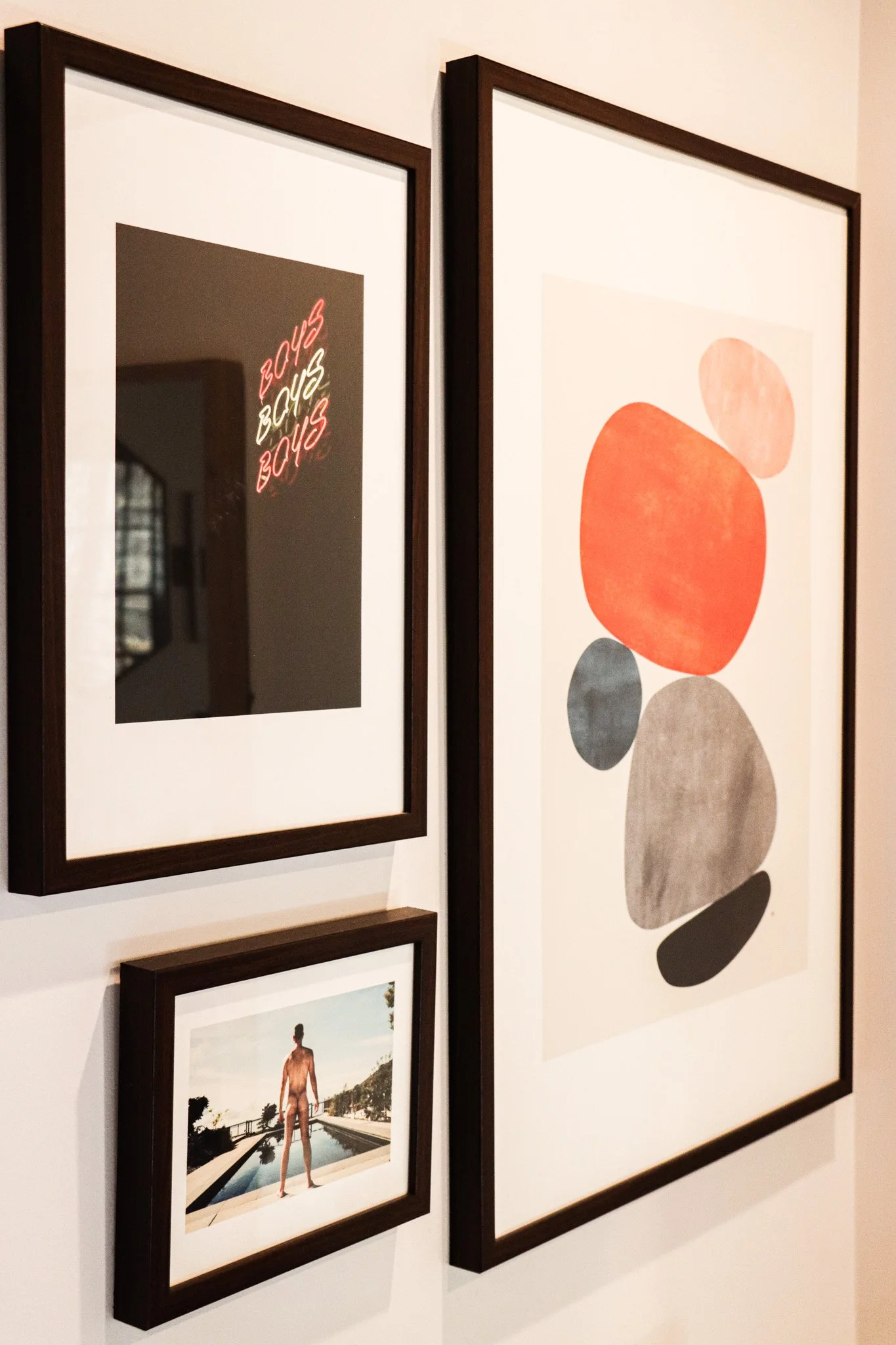A collage of framed art on a wall, including a neon sign with the word 'BOYS', an abstract artwork with colorful ovals, and a photograph of a person standing by a pool.
