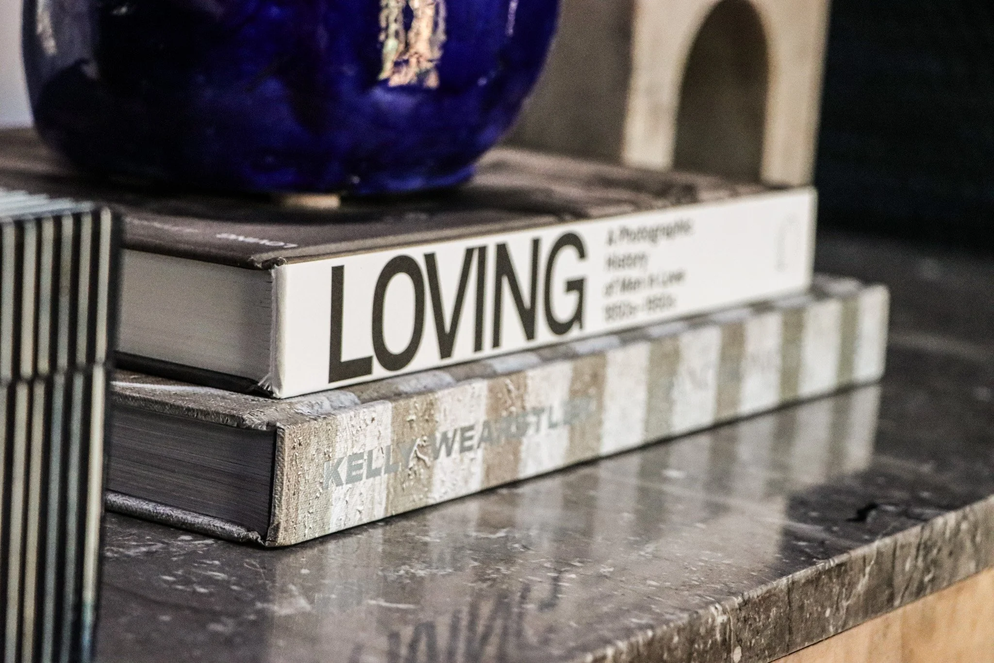 Close-up of two stacked books on a wooden surface, with a decorative purple and gold vase on top. The visible book titles are 'LOVING' and 'KELLY WELLS'.