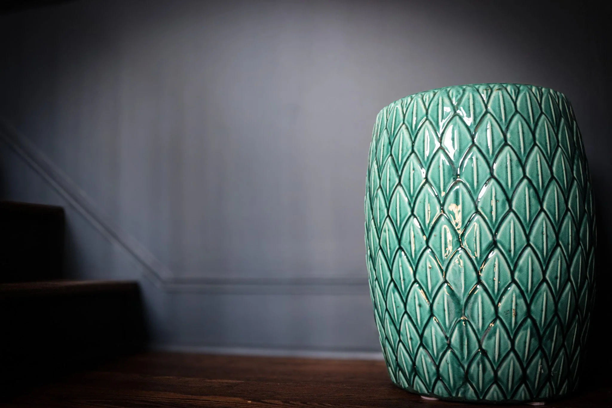 Close-up of a green ceramic vase with a textured, scale-like pattern, placed on a wooden surface near a staircase in a dimly lit interior.