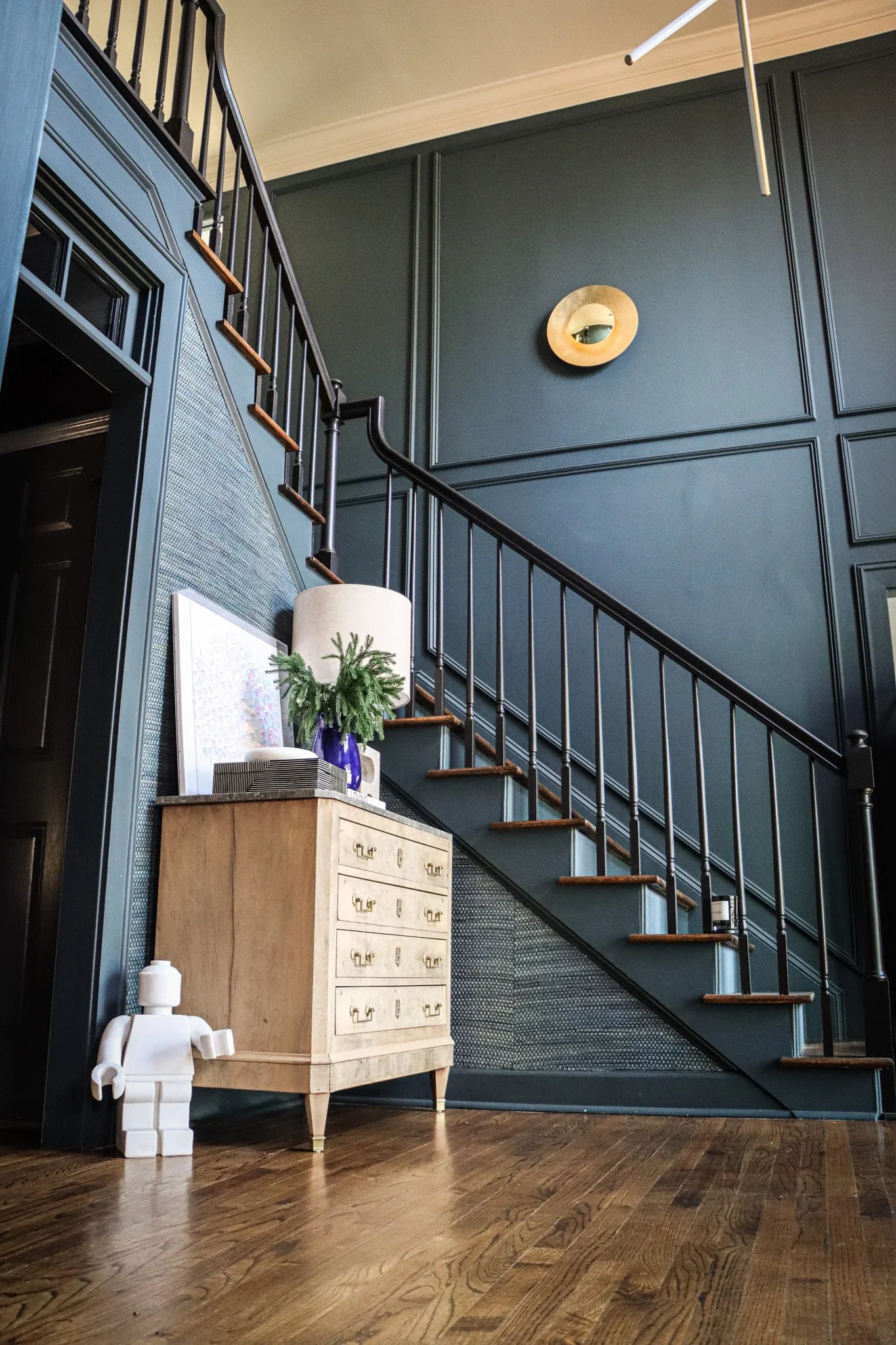 Interior view of a home with a staircase, dark green panel wall, a beige dresser with brass handles, a white Lego figure, a blue vase with greenery, and a lamp, with part of the ceiling and a circular mirror visible.