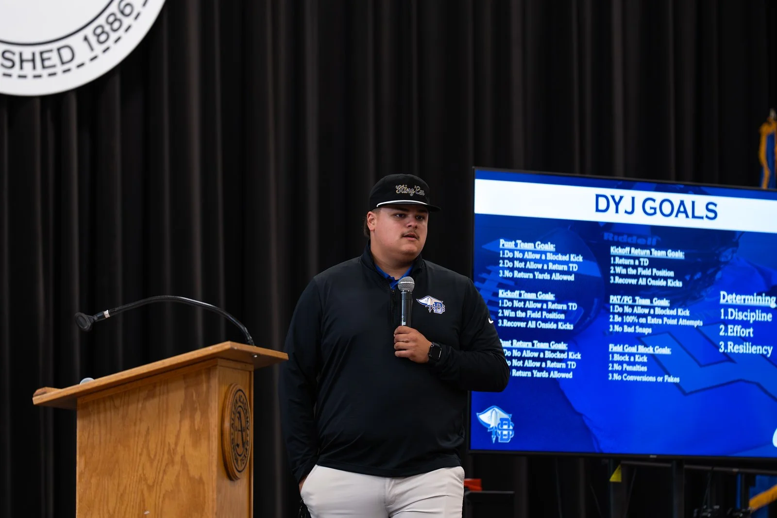 A young man stands with a microphone in hand, wearing a black cap and jacket, in front of a large digital screen displaying football goals and rules at an indoor event or presentation.