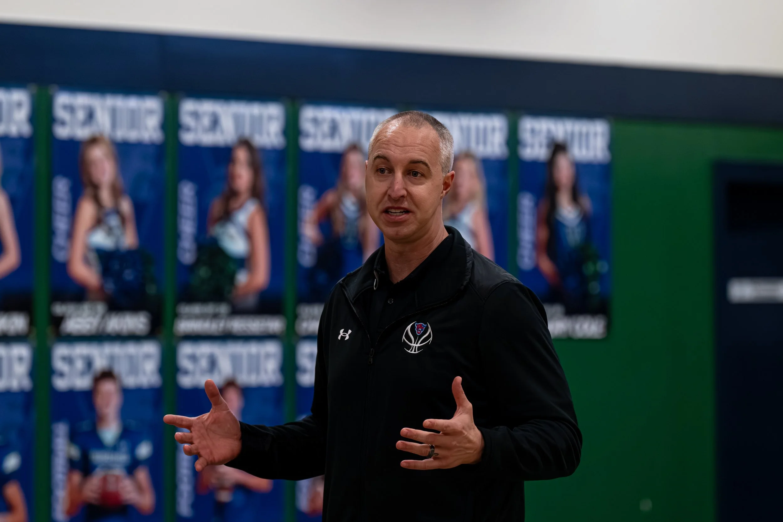 A man in a black jacket with a sports logo, speaking or giving instructions, standing in front of a wall with blue posters featuring women in cheerleading outfits.