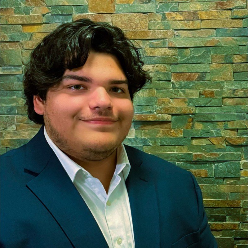 A young man with dark, wavy hair wearing a white dress shirt and navy blazer, smiling in front of a stone wall background.