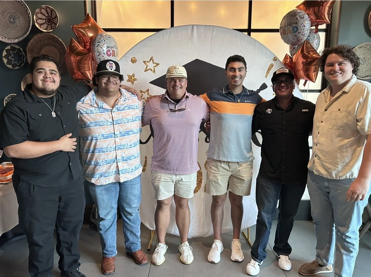 Group of six friends celebrating graduation, standing with arms around each other, with balloons and graduation-themed decorations in the background.