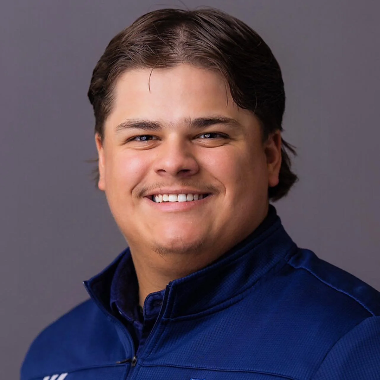 Portrait of a young man with dark hair smiling, wearing a blue sports jacket, against a gray background.