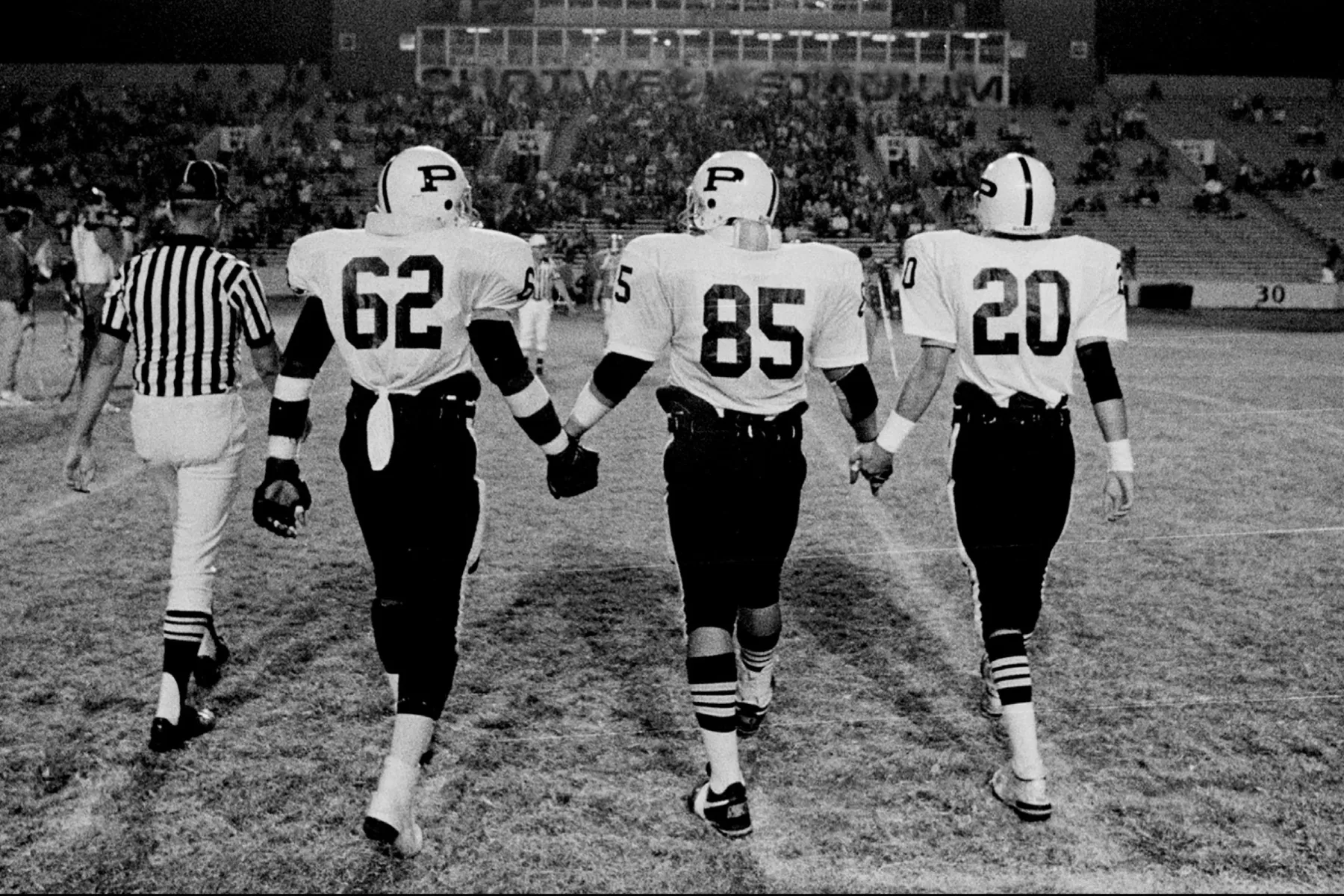 Three football players walking on the field holding hands, with a referee on the left, and stadium seats filled with spectators in the background.