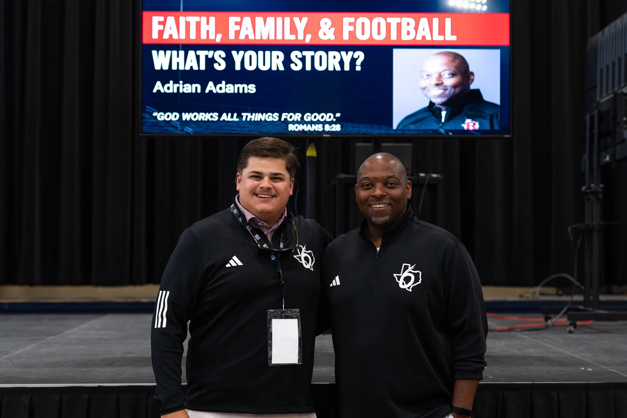 Two men standing together indoors, wearing black tracksuits with a logo, smiling for the camera. Behind them is a large screen displaying a presentation titled 'Faith, Family, & Football' featuring a photo and name of Adrian Adams, with a quote from Romans 8:28.