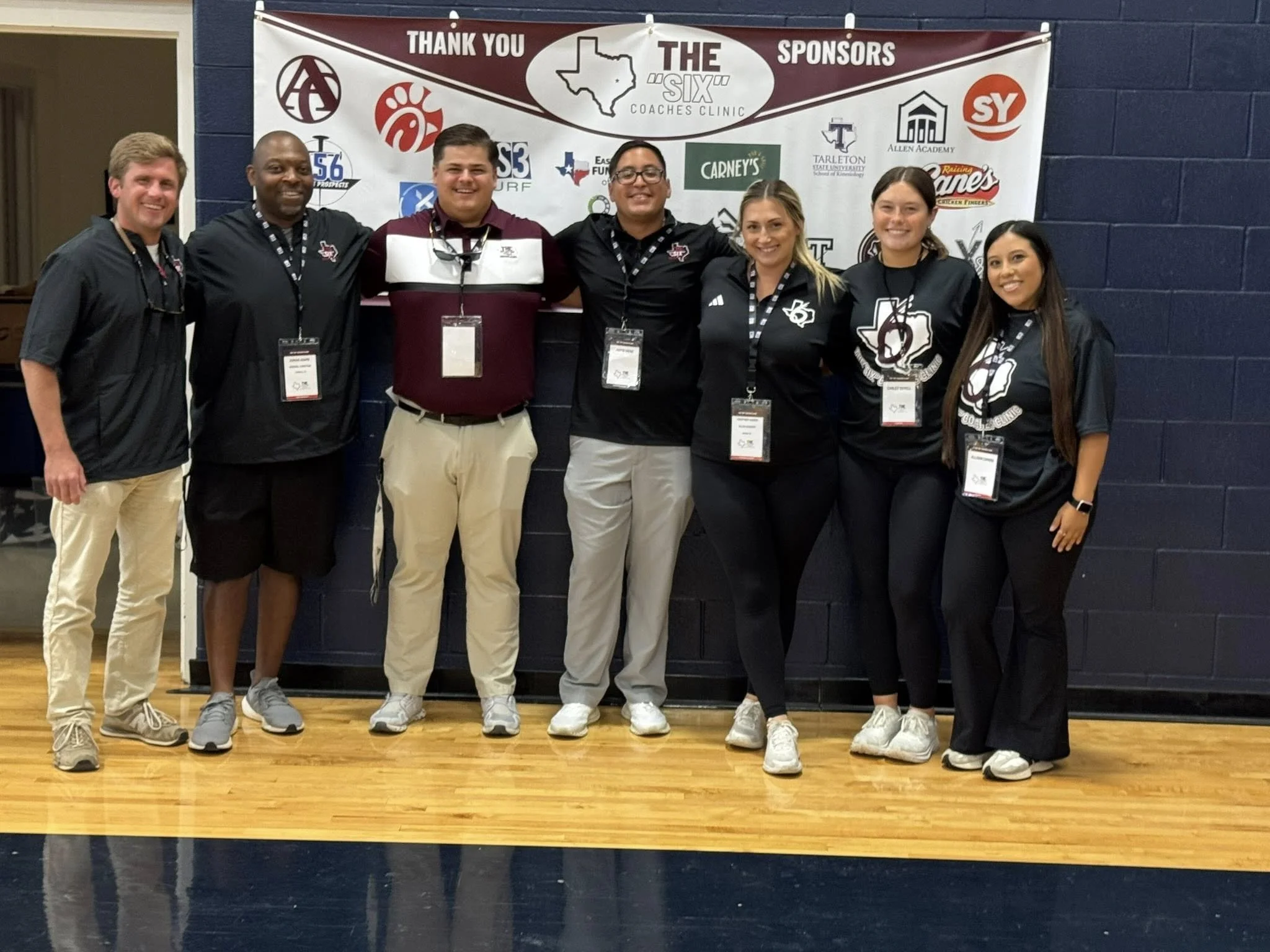 Group of seven people standing in front of a sponsor banner at a school indoor event, smiling at the camera.