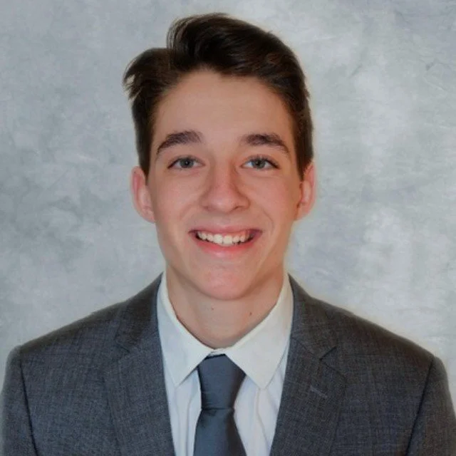 Young man in a gray suit, white shirt, and gray tie smiling in front of a plain background.