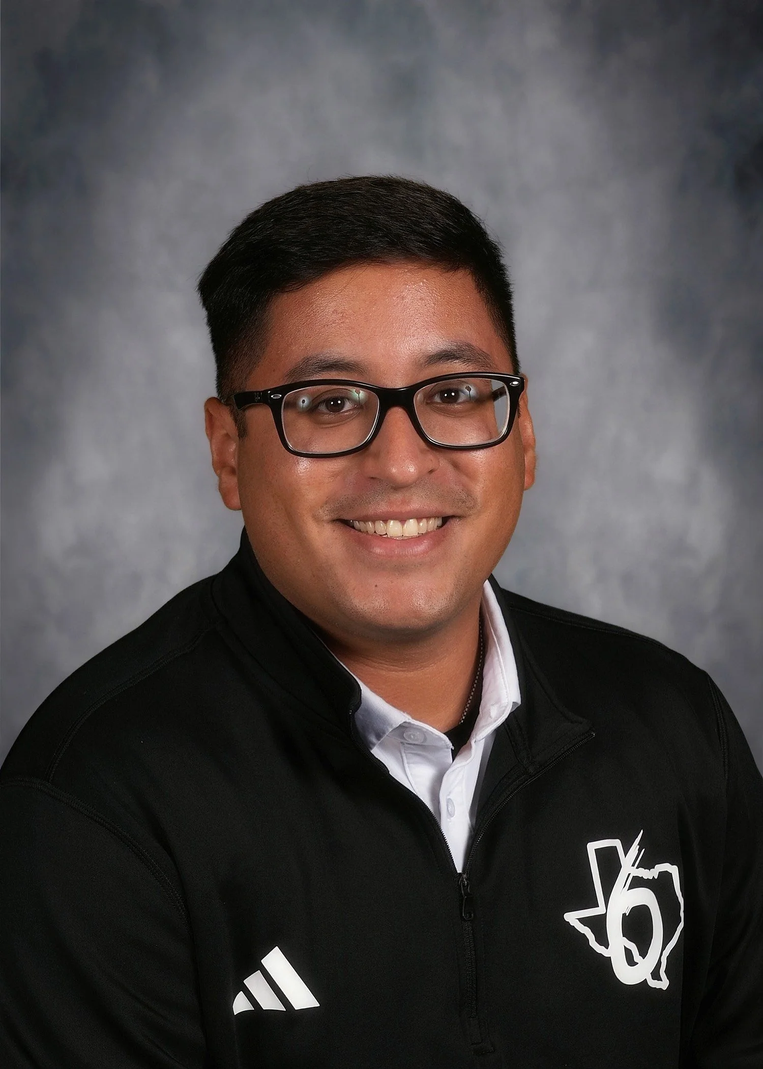 A young man with black hair, wearing glasses, a black zip-up jacket with a white logo, and a white collared shirt, smiling against a gray textured background.