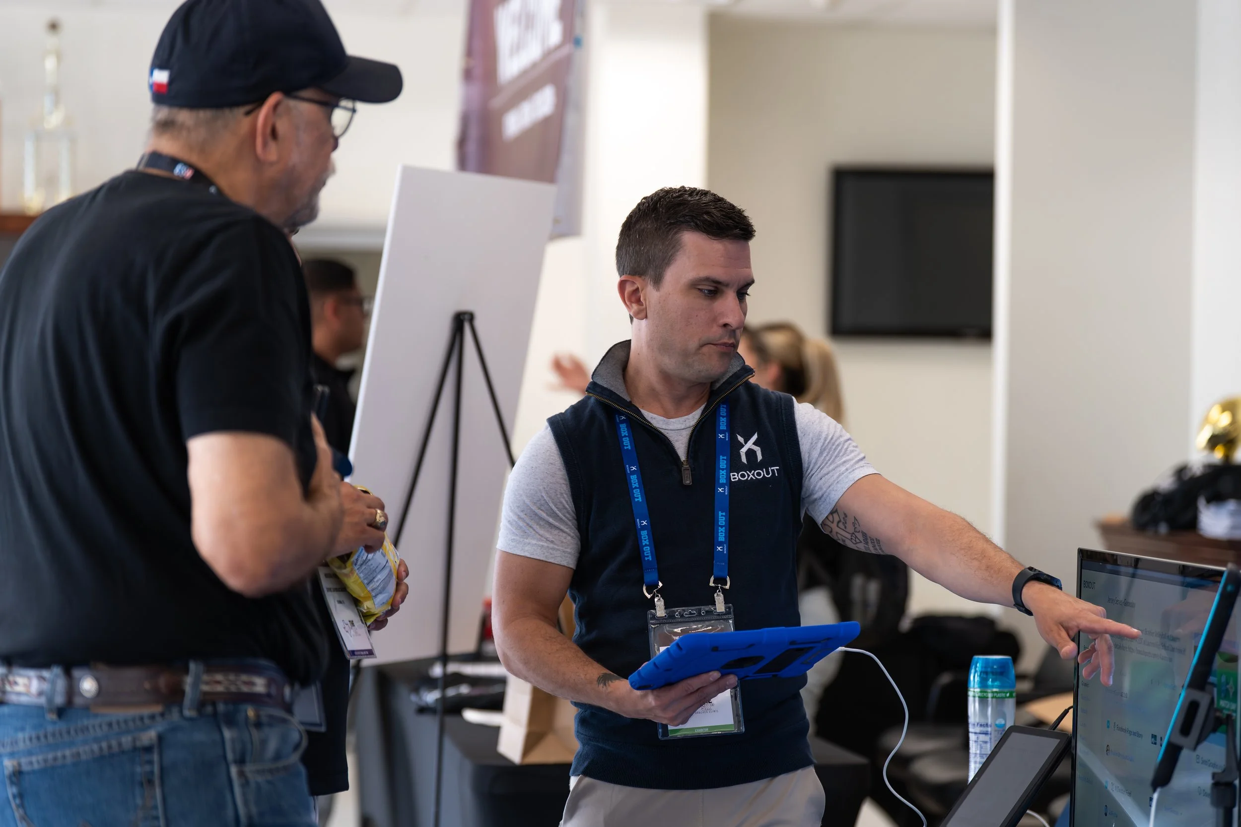 Two men at a conference or trade show. One man, wearing a black cap and shirt, is holding snacks. The other man, wearing a gray shirt with a black vest, is interacting with a screen and holding a tablet. There are other people and a whiteboard in the background.