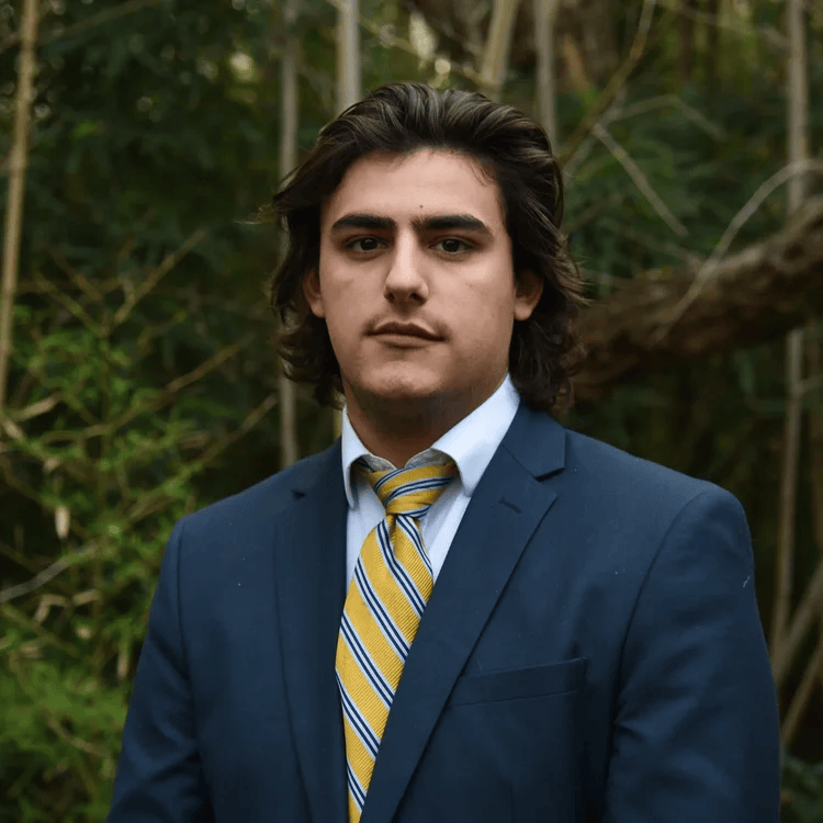 Young man in a navy suit and yellow striped tie standing outdoors with trees and green foliage in the background.