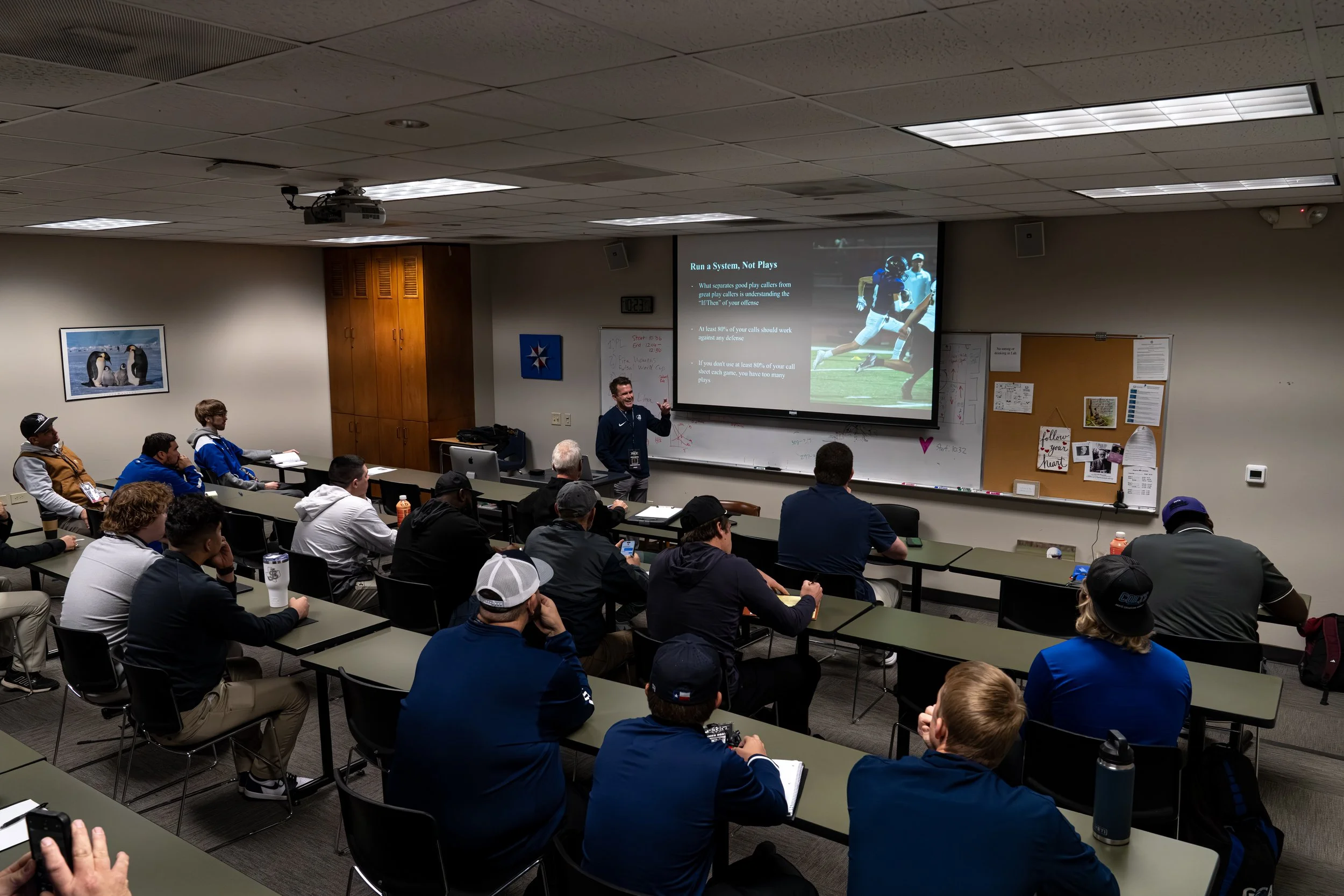 A classroom filled with students attending a lecture. A instructor stands in front of a projection screen displaying a presentation slide about football strategy. The walls are decorated with photos and posters, and students are seated at long tables, some taking notes, listening attentively.