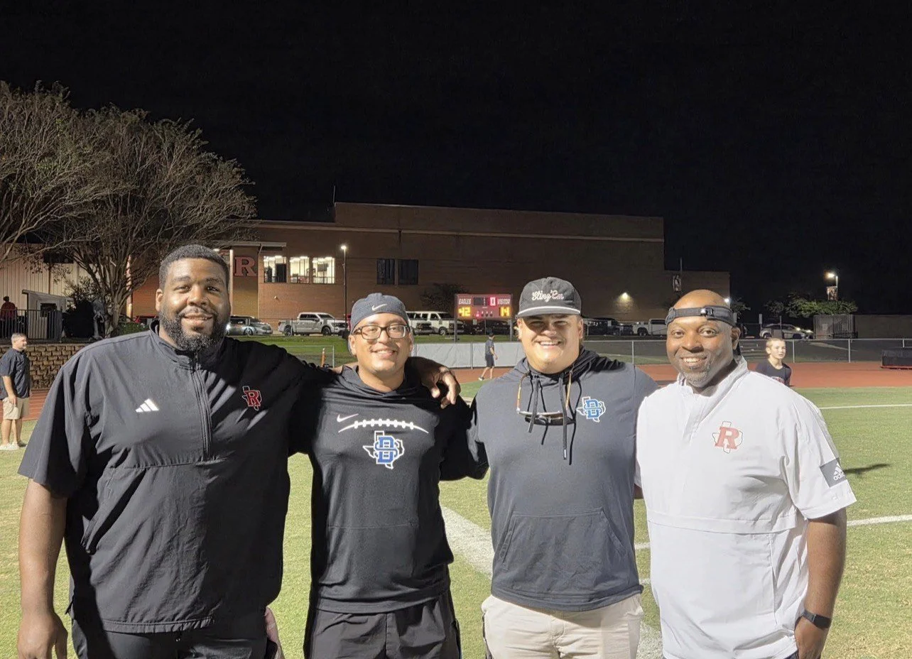 Four men standing together on a football field at night, smiling for the camera. They are wearing sports apparel with logos, indicating they are likely coaches or team staff.