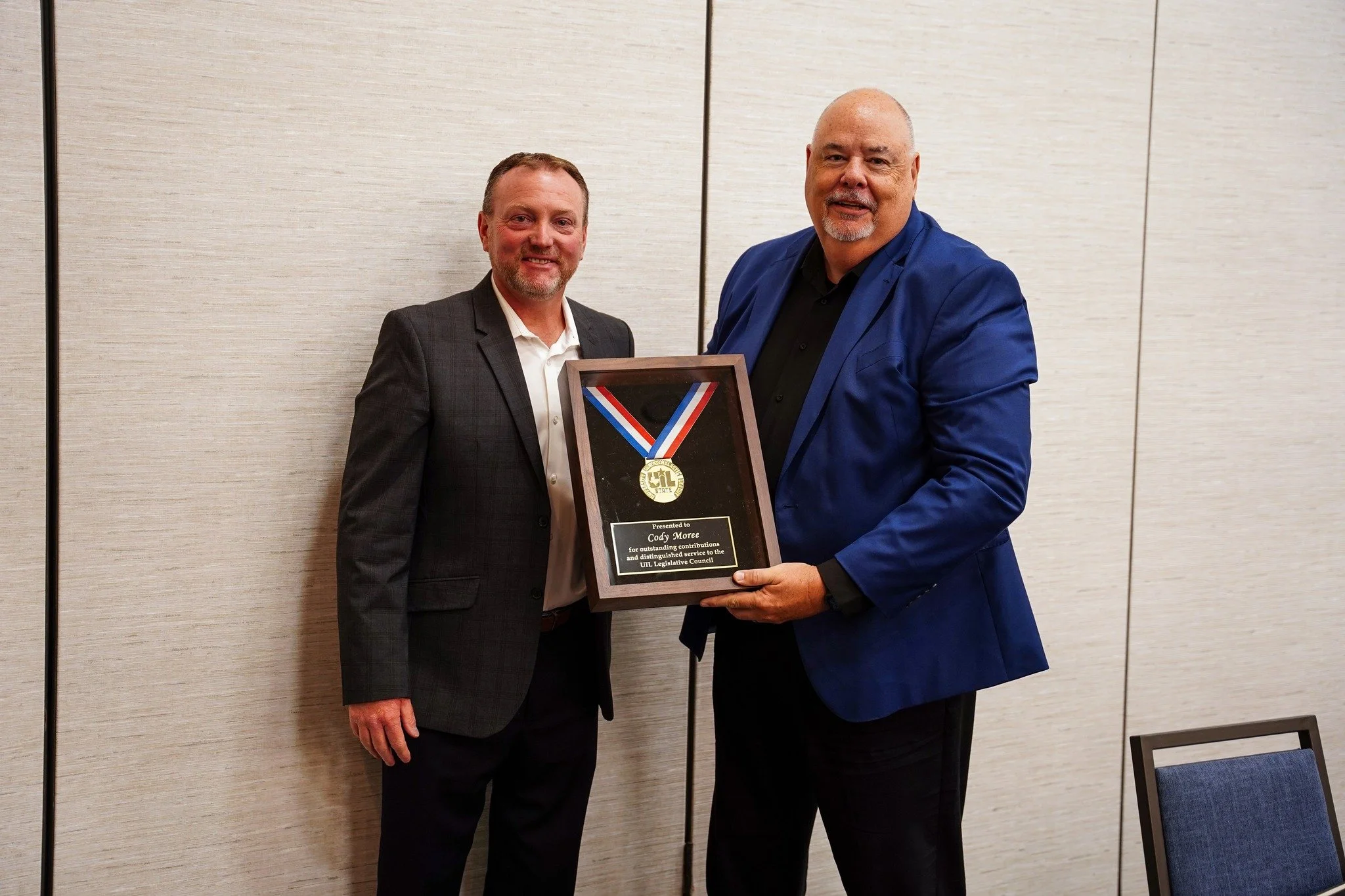 Two men standing next to each other, holding a framed award with a medal hanging above it. The man on the left is wearing a gray suit with a white shirt, and the man on the right is wearing a blue jacket and black shirt.