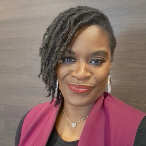 A woman with styled dreadlocks, wearing a maroon blazer over a black top, silver earrings, and a heart-shaped necklace, smiling in front of a neutral background.