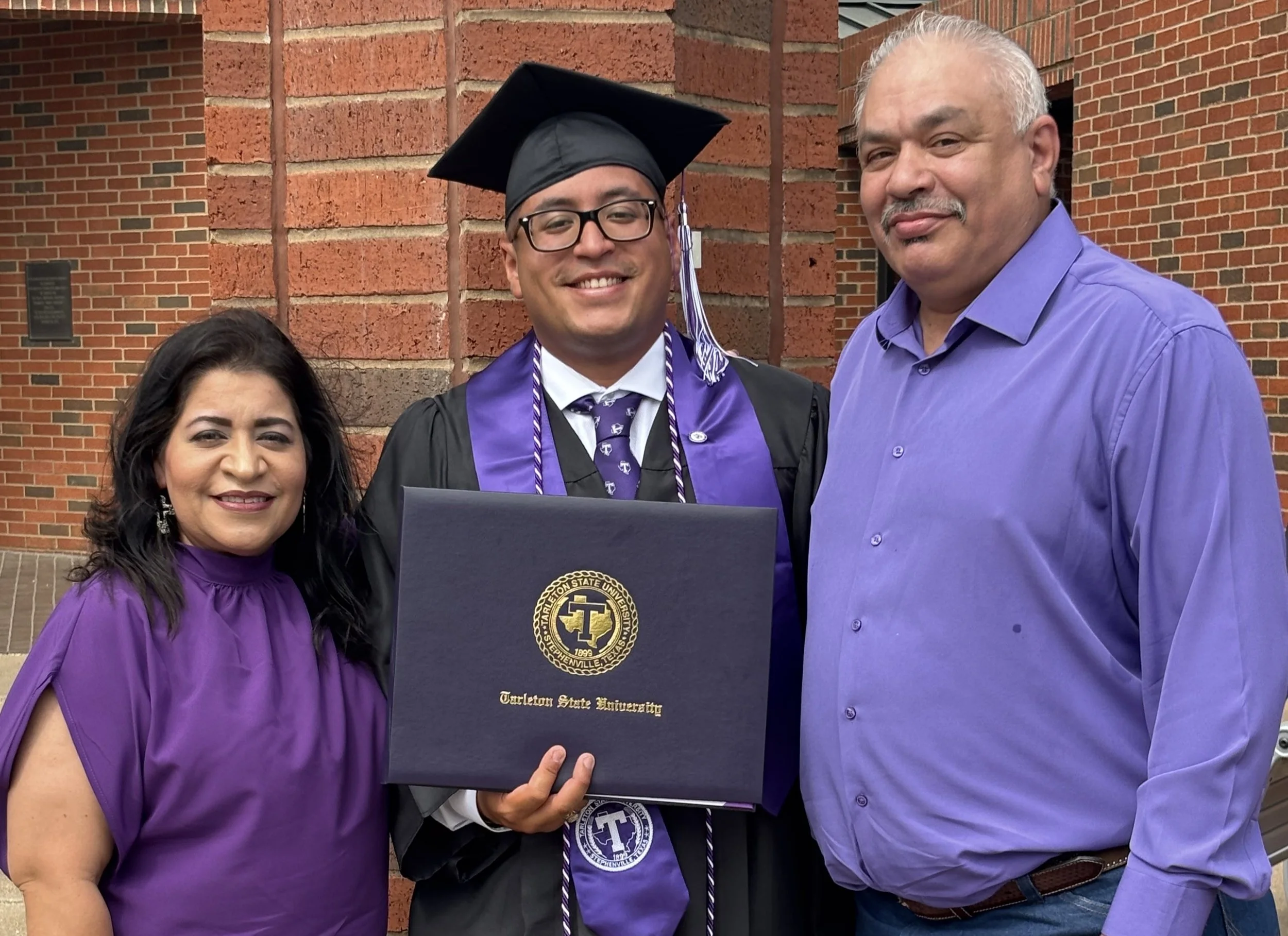 A young man in a graduation cap and gown holding a diploma, standing with a woman in a purple dress and a man in a purple shirt, outside a brick building.