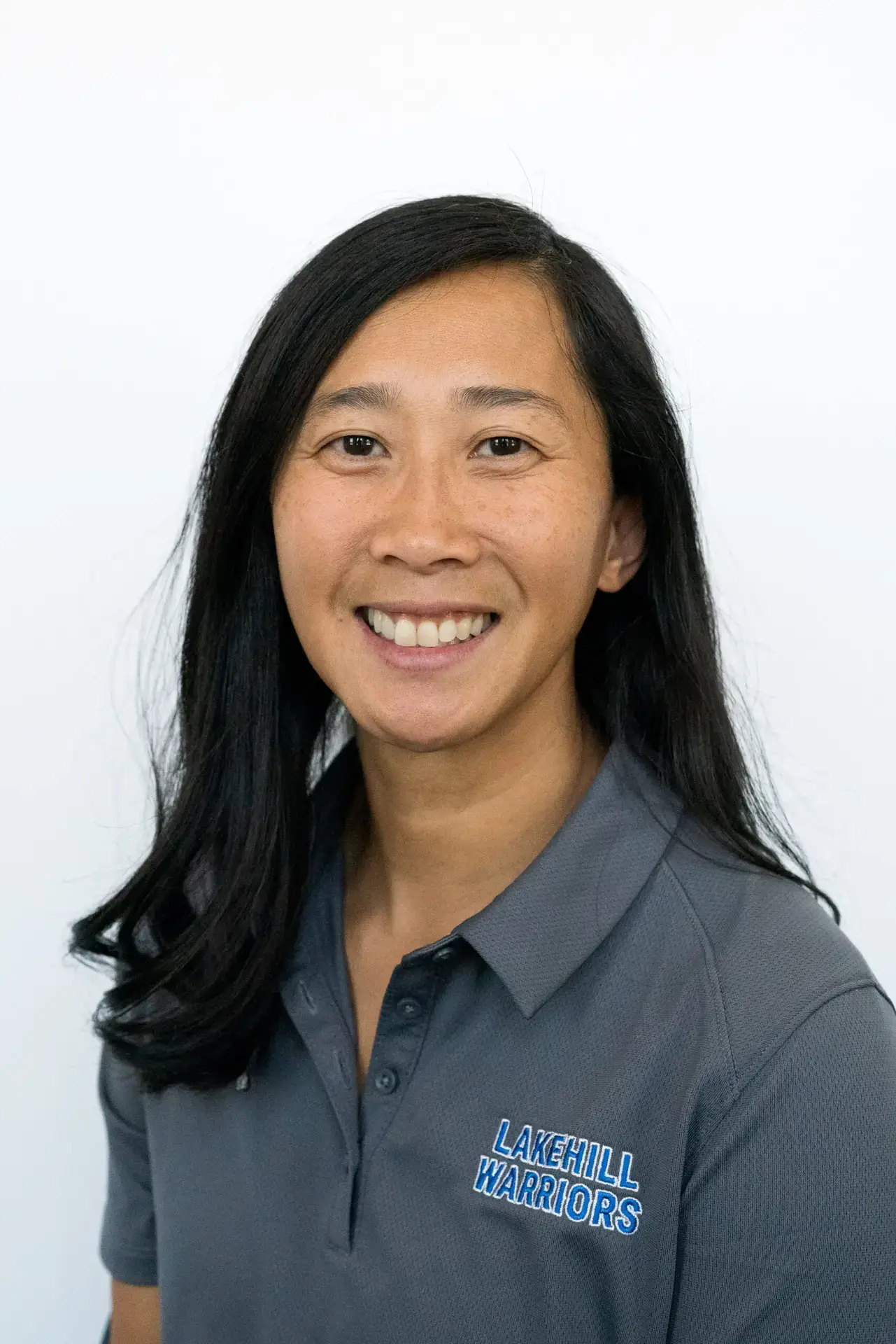 A woman with long black hair smiling, wearing a gray polo shirt with "Lakehills Warriors" embroidered on it, against a plain white background.