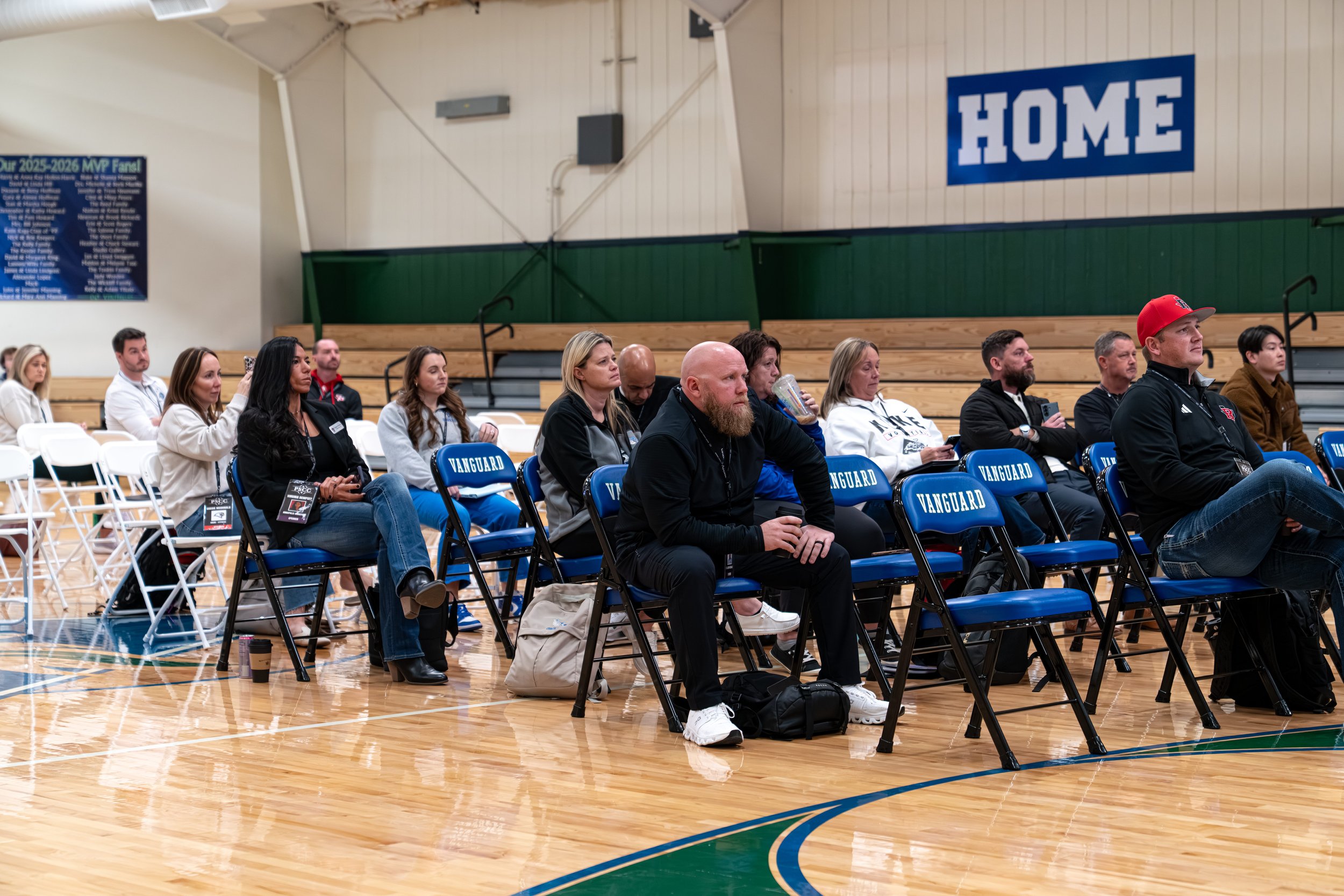 People sitting on chairs in an indoor gymnasium, attending an event or presentation. The chairs are blue with 'VANGUARD' written on the back. The audience appears focused, some taking notes or drinking from cups. The gym has a wooden floor, green and white walls, and a large blue sign with the word 'HOME' on the wall.