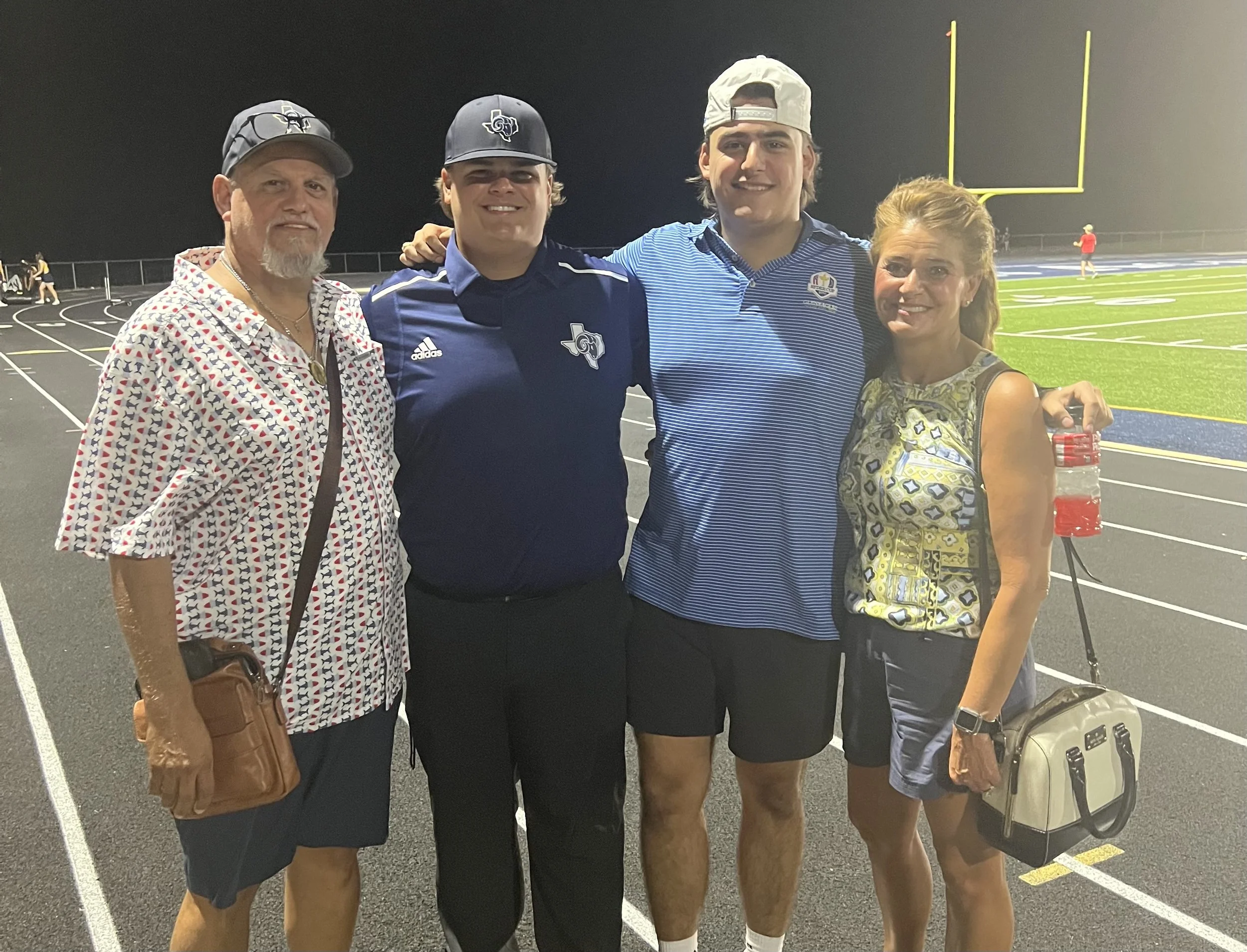 Four people standing on a football field track at night, smiling for the camera. Two men are wearing sports attire with football team logos, and two women are dressed casually, one with a purse and the other with a small bag. The background shows football goal posts and a field, with a person walking in the distance.