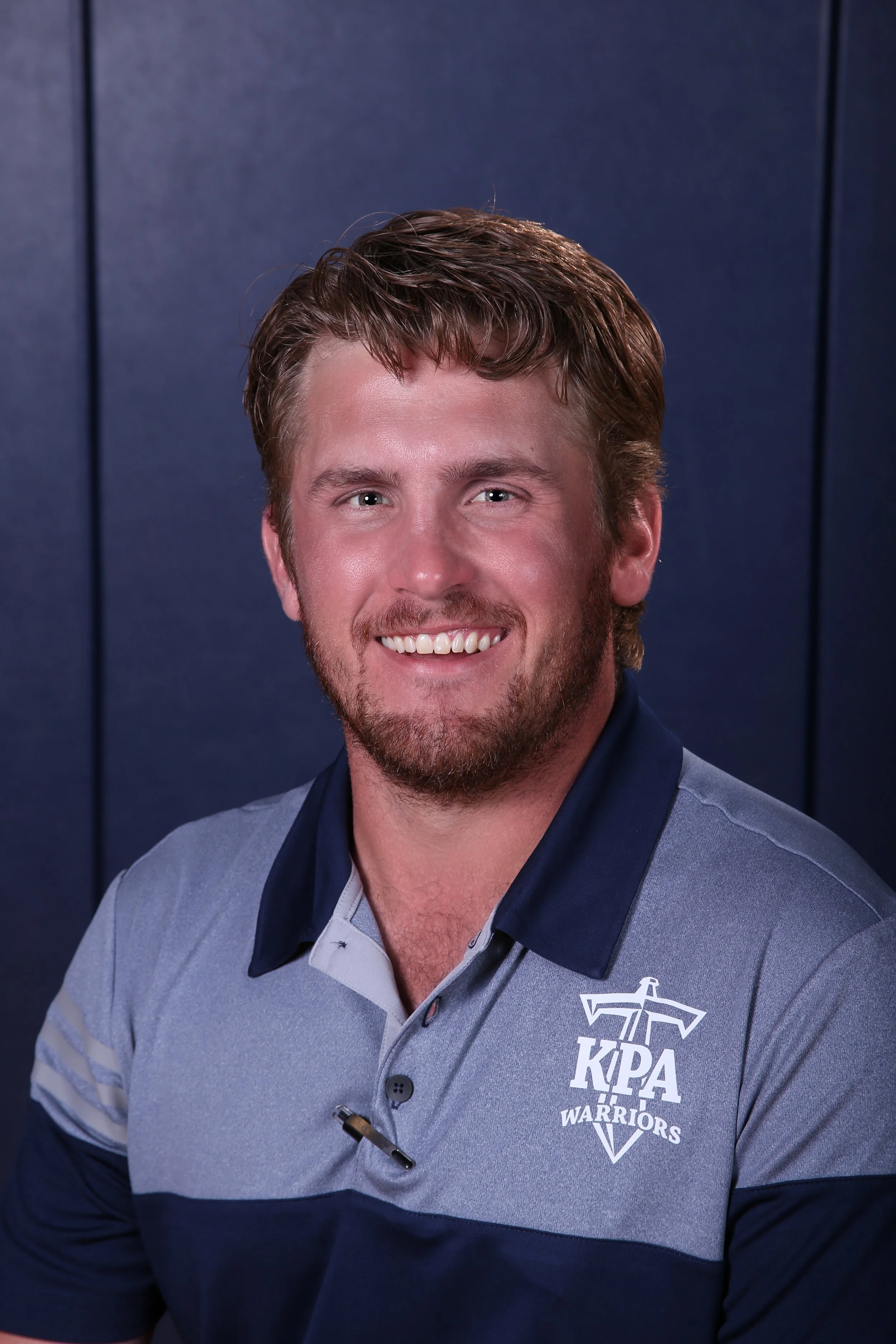 A man with brown hair and a beard smiling, wearing a gray and navy blue polo shirt with a KPAX Warriors logo.