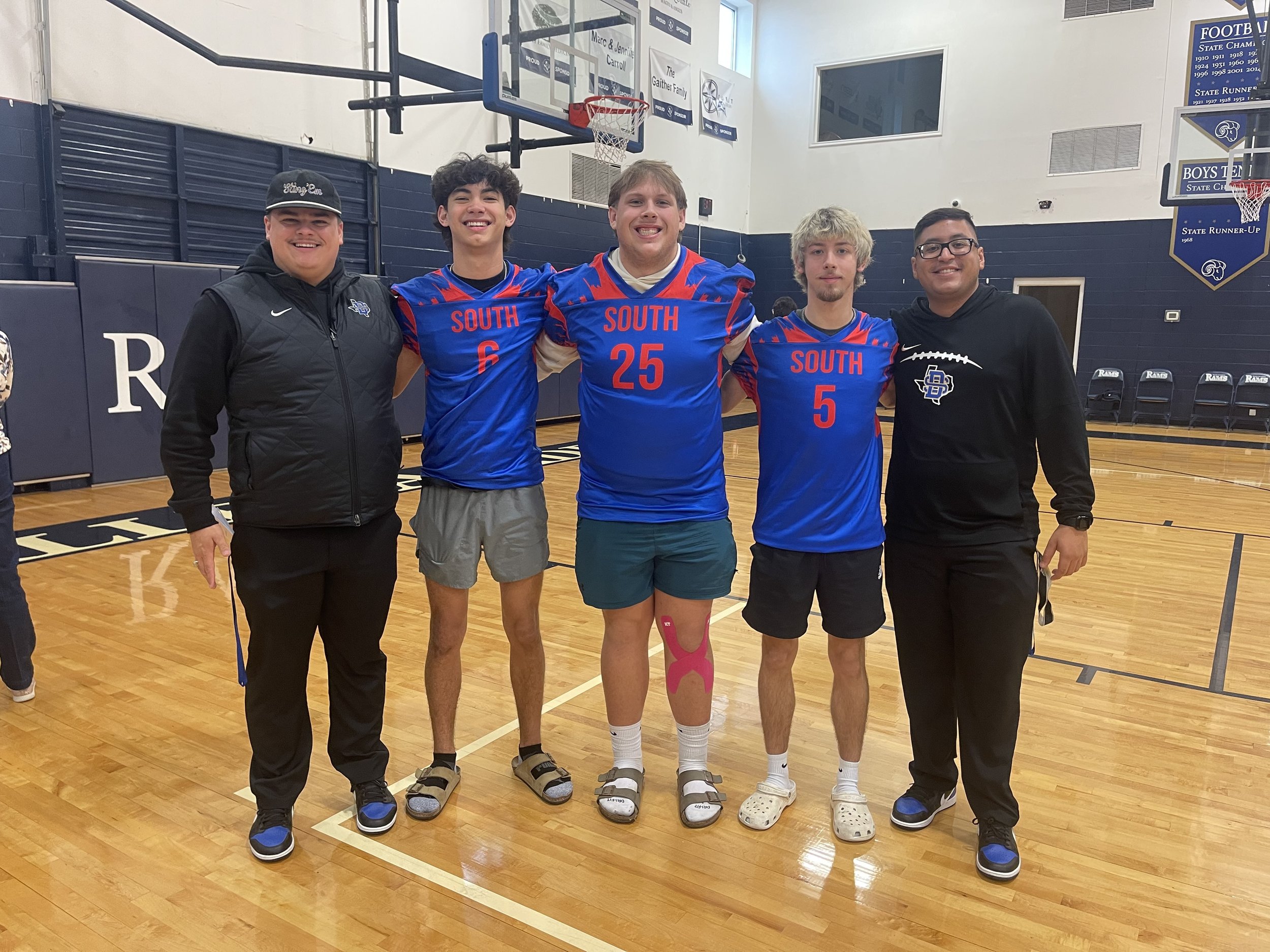 Five young men standing together in a gymnasium, smiling, with two wearing football uniforms labeled 'South' and three in casual sportswear.