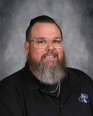 A man with a beard, glasses, and long hair, wearing a black polo shirt with a logo on it, posing for a portrait against a gray background.
