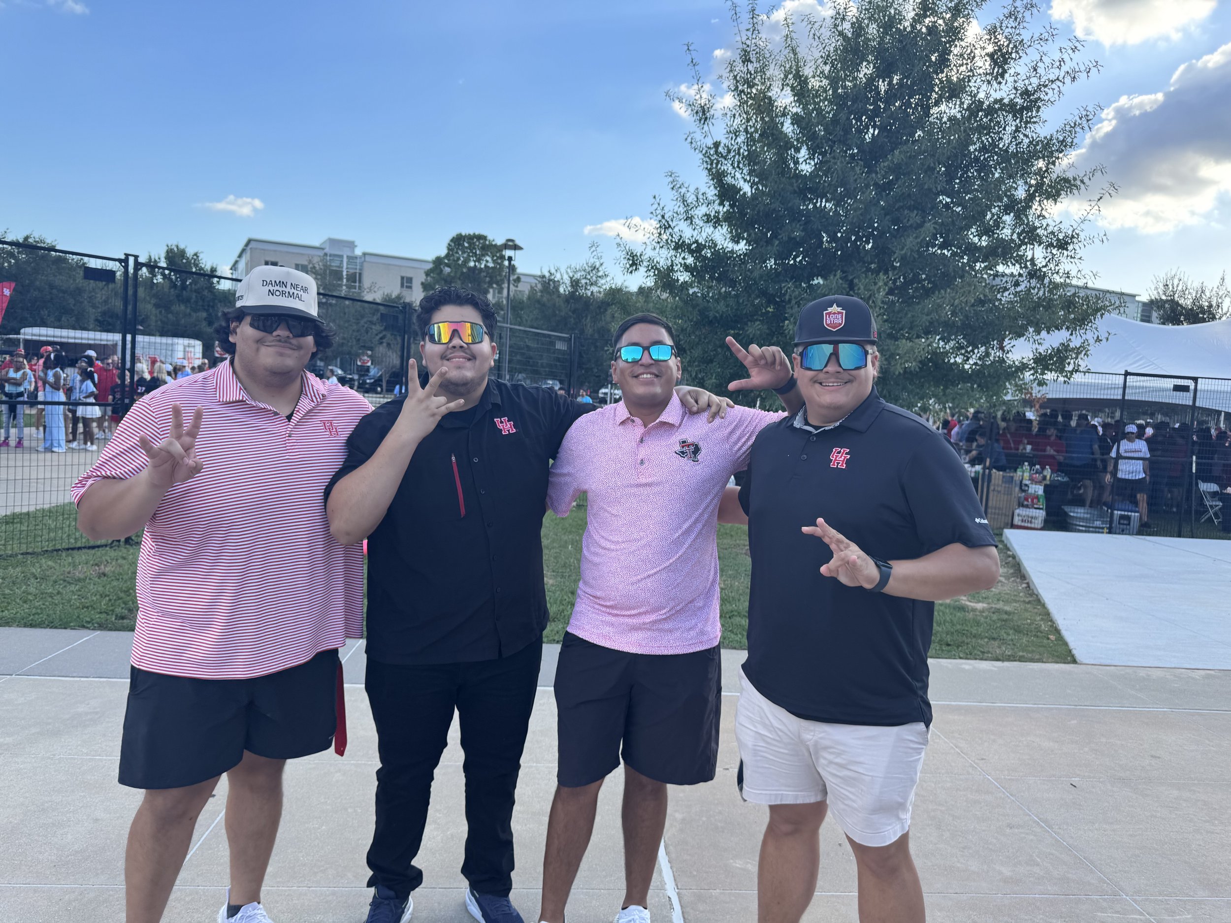 Four men standing together outdoors at a social gathering or event, smiling and posing for the photo, with people and tents in the background.
